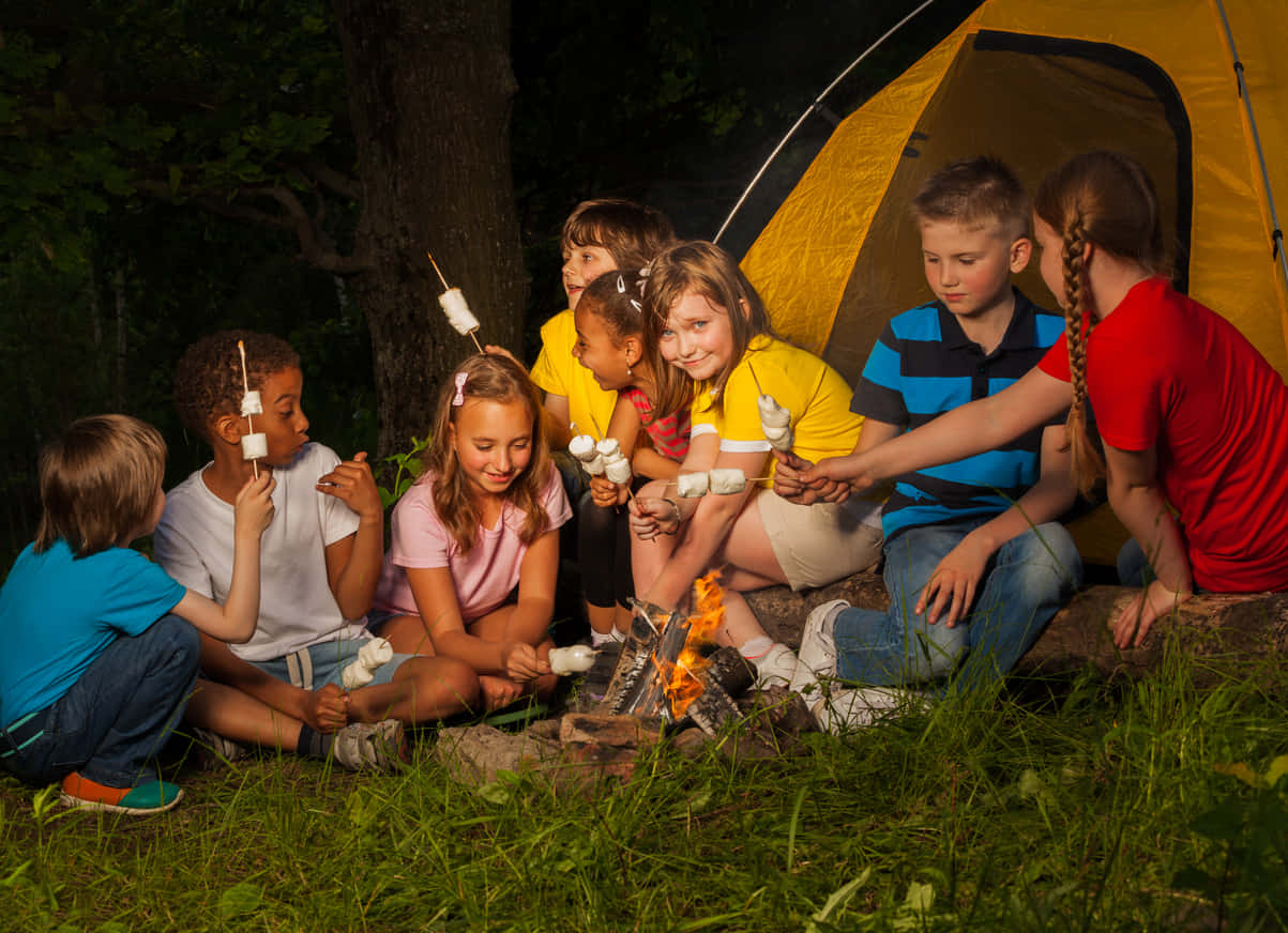 Camping 桌布 壁紙 — 孩子們在夜晚聚集在營火旁，在黃色帳篷附近的火焰光芒下烤棉花糖。