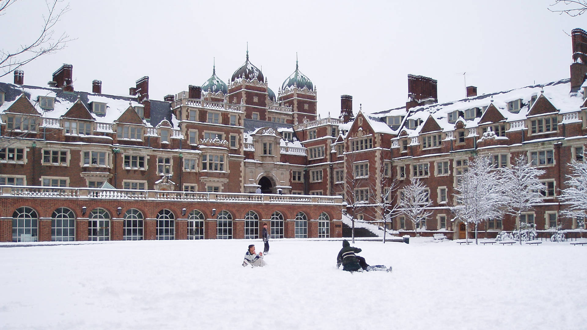 雪 桌布 壁紙 — 孩子們在宏偉的紅磚大學建築前，沿著積雪的山坡滑雪橇，周圍有積雪的樹木。