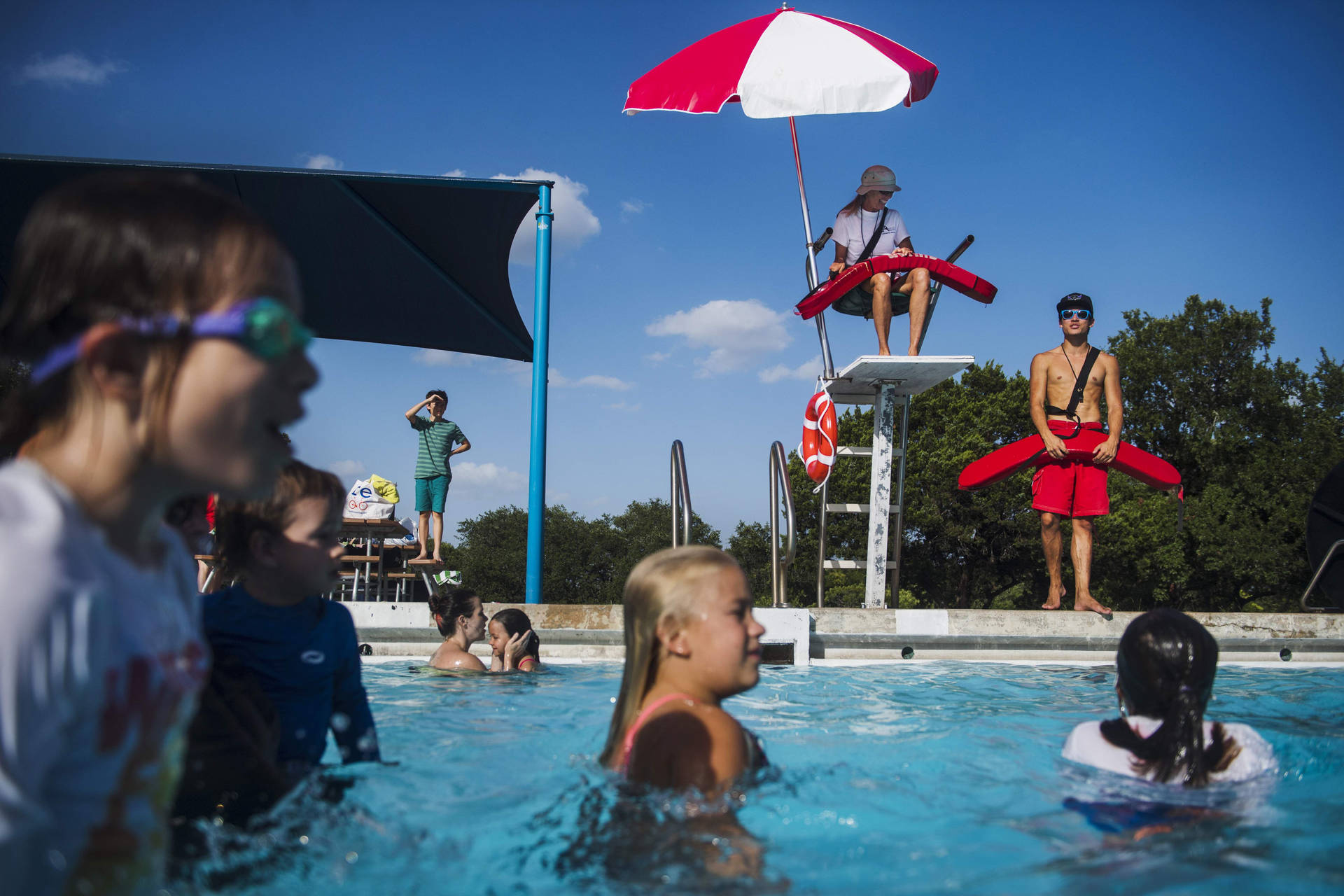 Swimming 桌布 壁紙 — 孩子們在晴朗的天空下，在明亮的藍色戶外泳池中游泳和玩耍，救生員從高椅上監視。