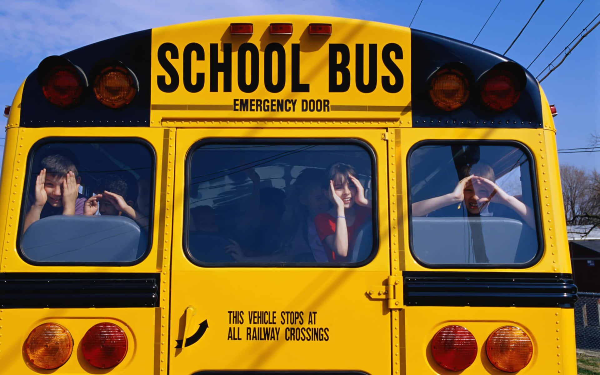 Yellow School Bus 桌布 壁紙 — 孩子們從明亮的黃色校車窗戶探出頭來，在晴朗的藍天下做出俏皮的手勢。