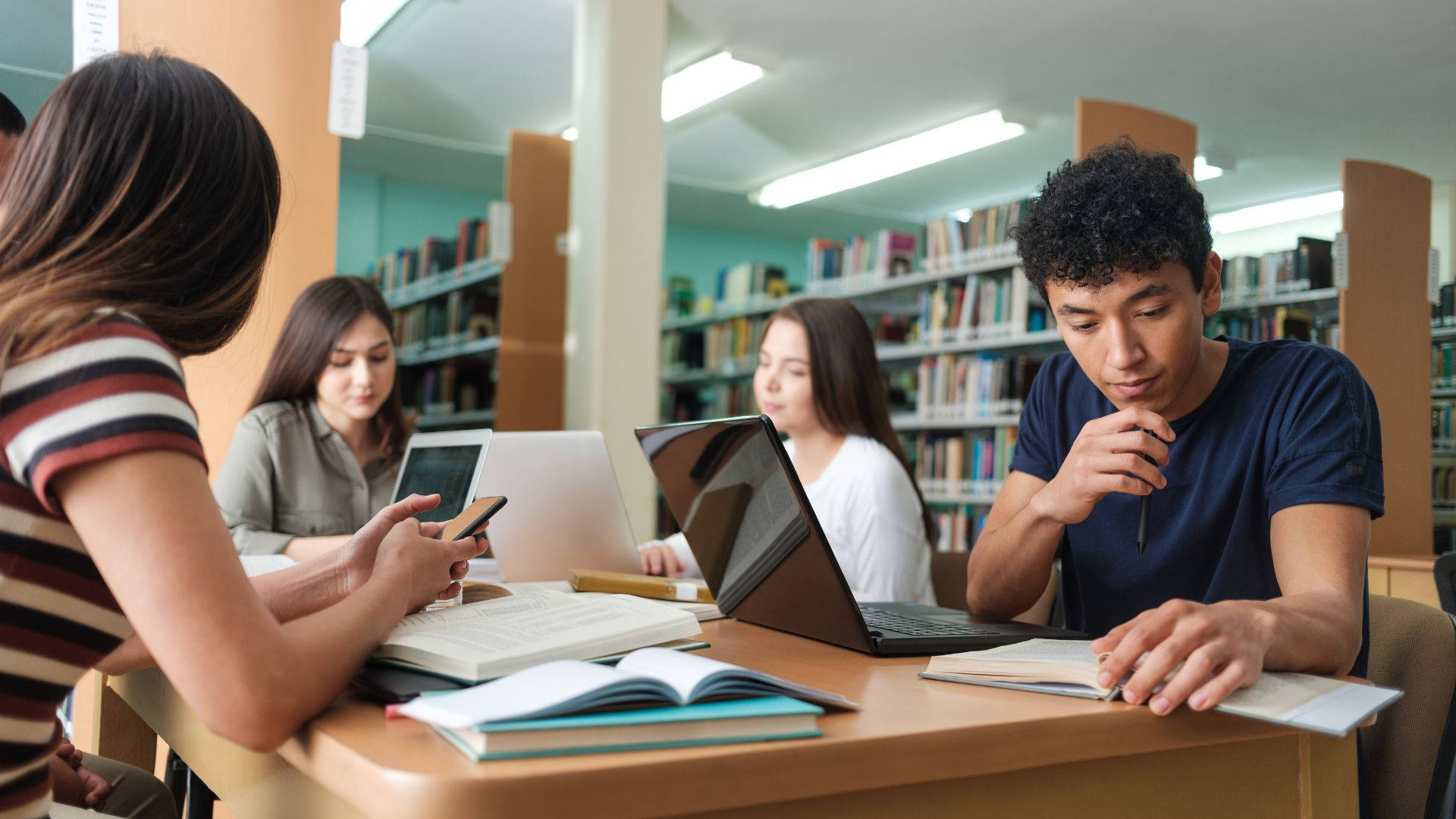 books 桌布 壁紙 — 學生們在明亮的圖書館裡，被書架和打開的筆記型電腦包圍著，專心學習。