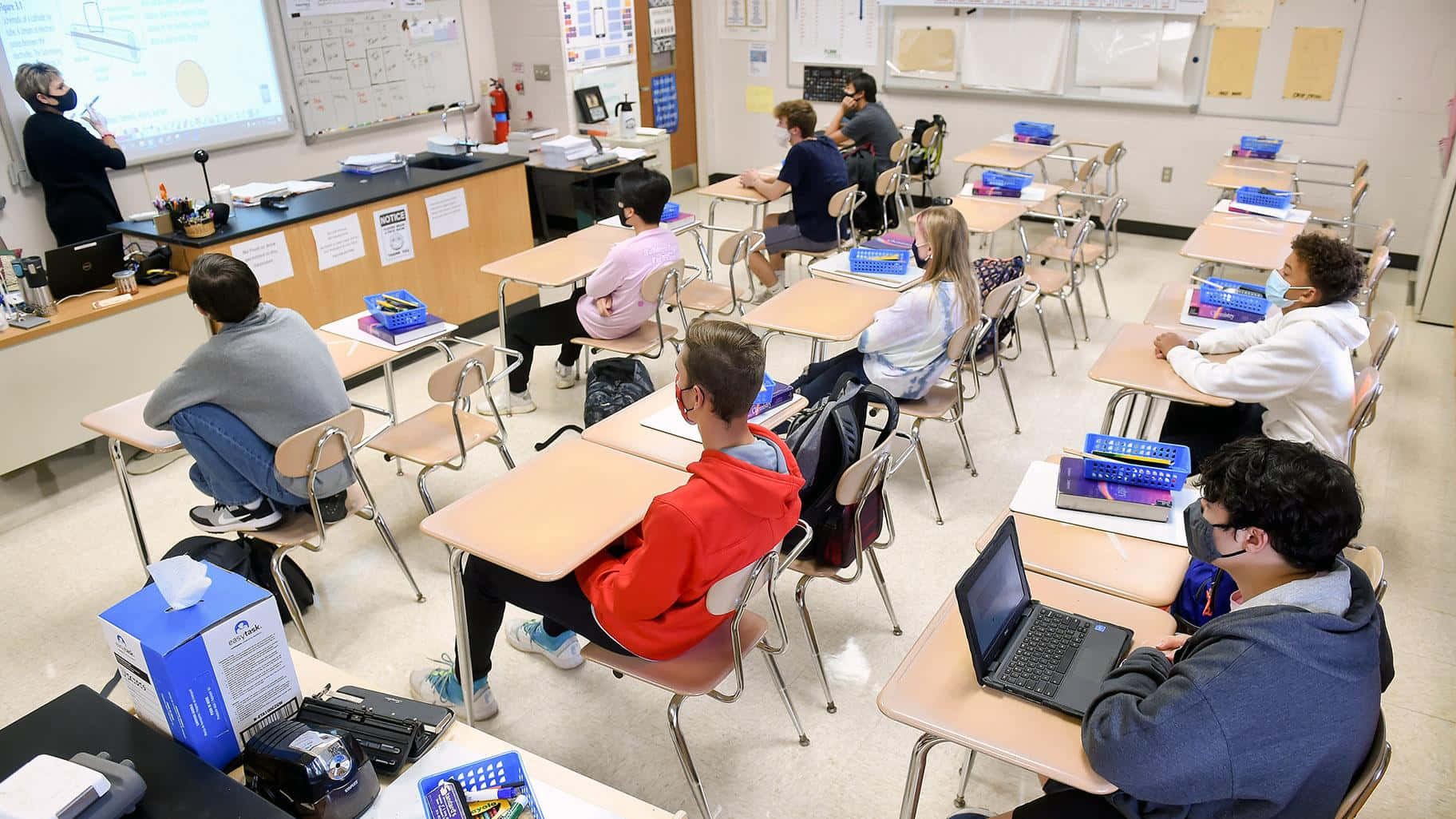 Classroom 桌布 壁紙 — 學生戴著口罩坐在教室的書桌前，老師在前方靠近白板和投影機的位置。