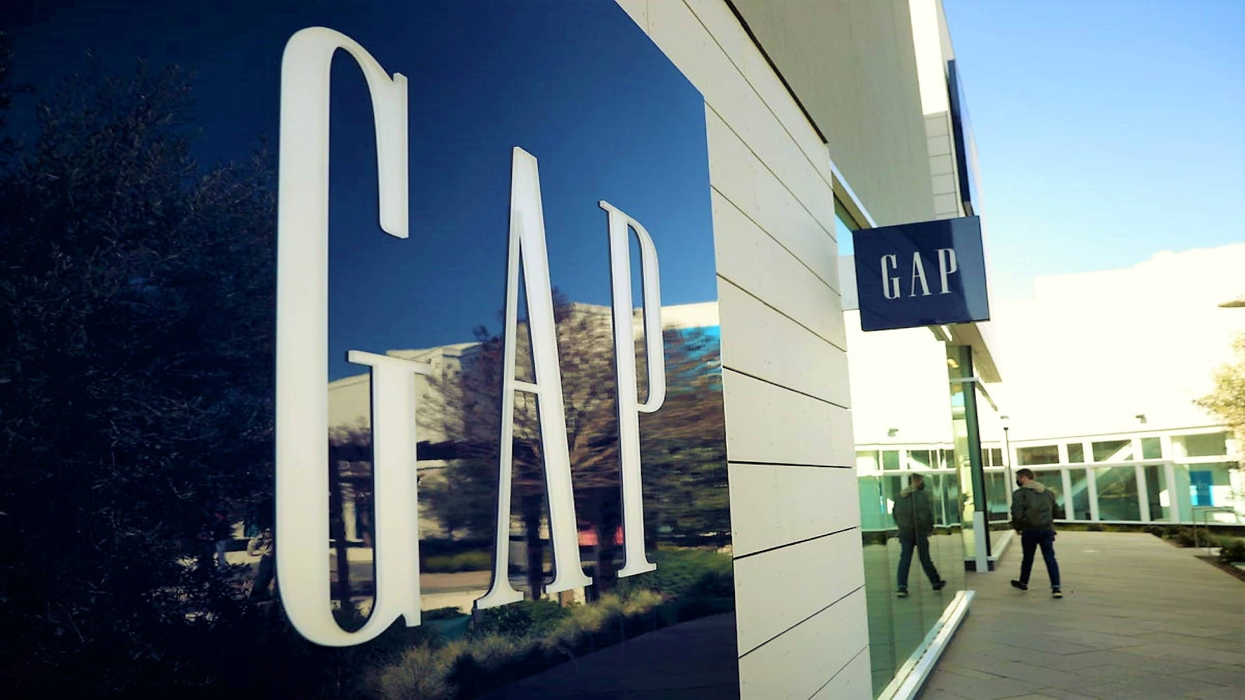 Gap 桌布 壁紙 — 安裝在現代建築外牆上的大型白色GAP字母，在明亮的陽光下反射出清澈的藍天和樹木。
