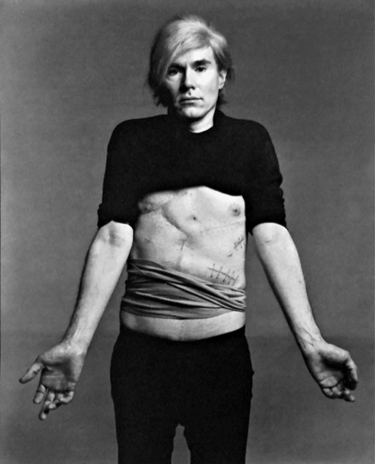 andy warhol 桌布 壁紙 — 安迪·沃荷站在簡陋的工作室，身穿破爛襯衫，露出縫合的腹部疤痕，背景是純灰色。
