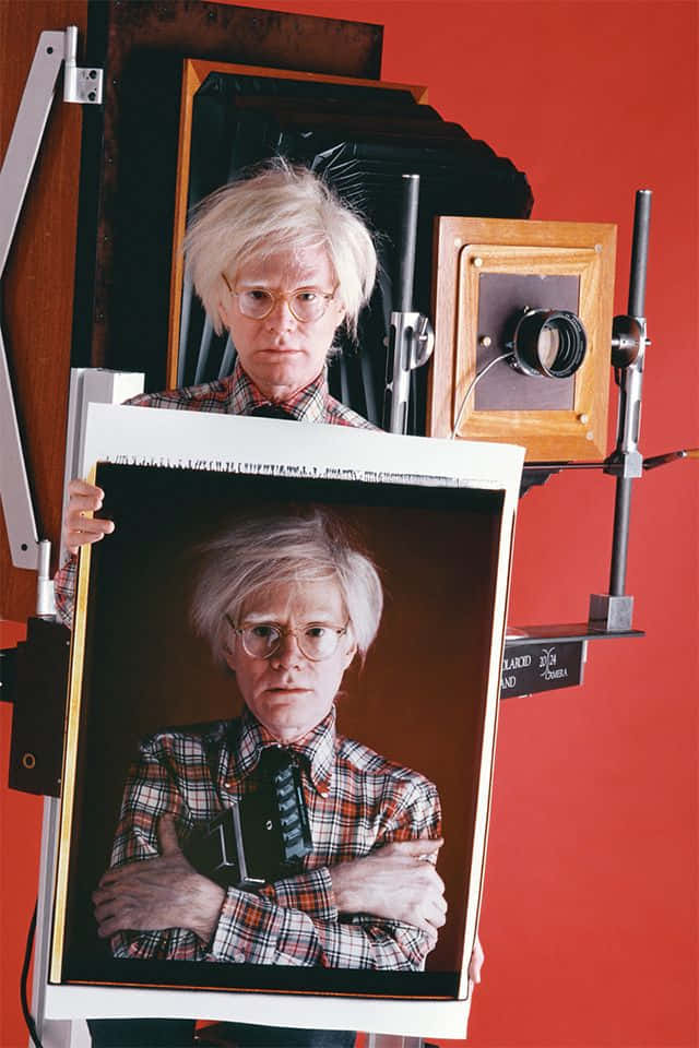 andy warhol 桌布 壁紙 — 安迪·沃荷站在自己拿著相機的大型照片印刷品後面，背景是鮮紅色的牆壁和復古攝影器材。