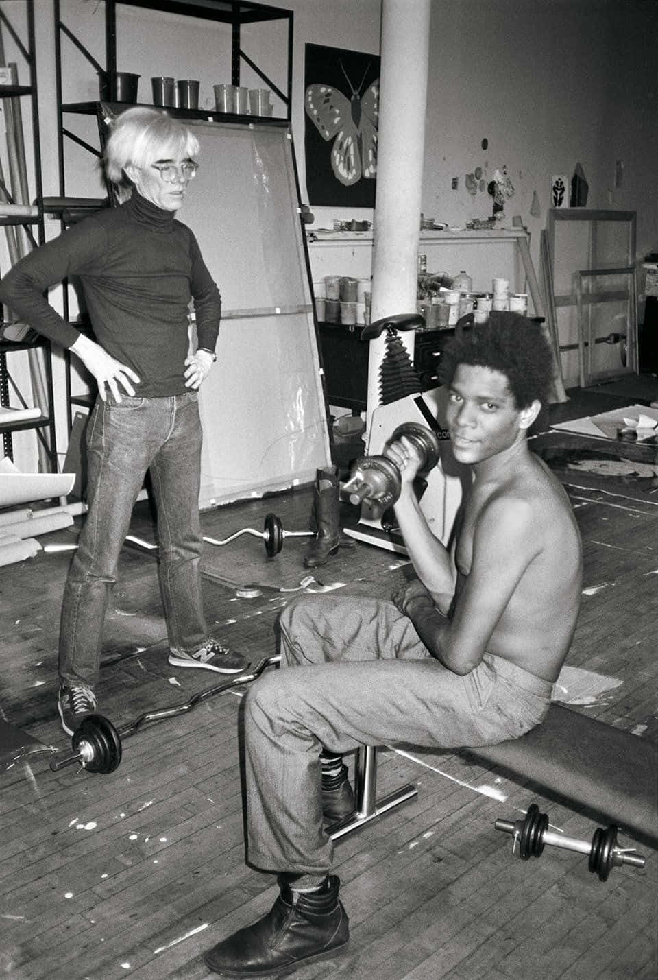 andy warhol 桌布 壁紙 — 安迪·沃荷雙手叉腰站立，一名赤裸上身的男子在堆滿美術用品和啞鈴的凌亂工作室中舉重。