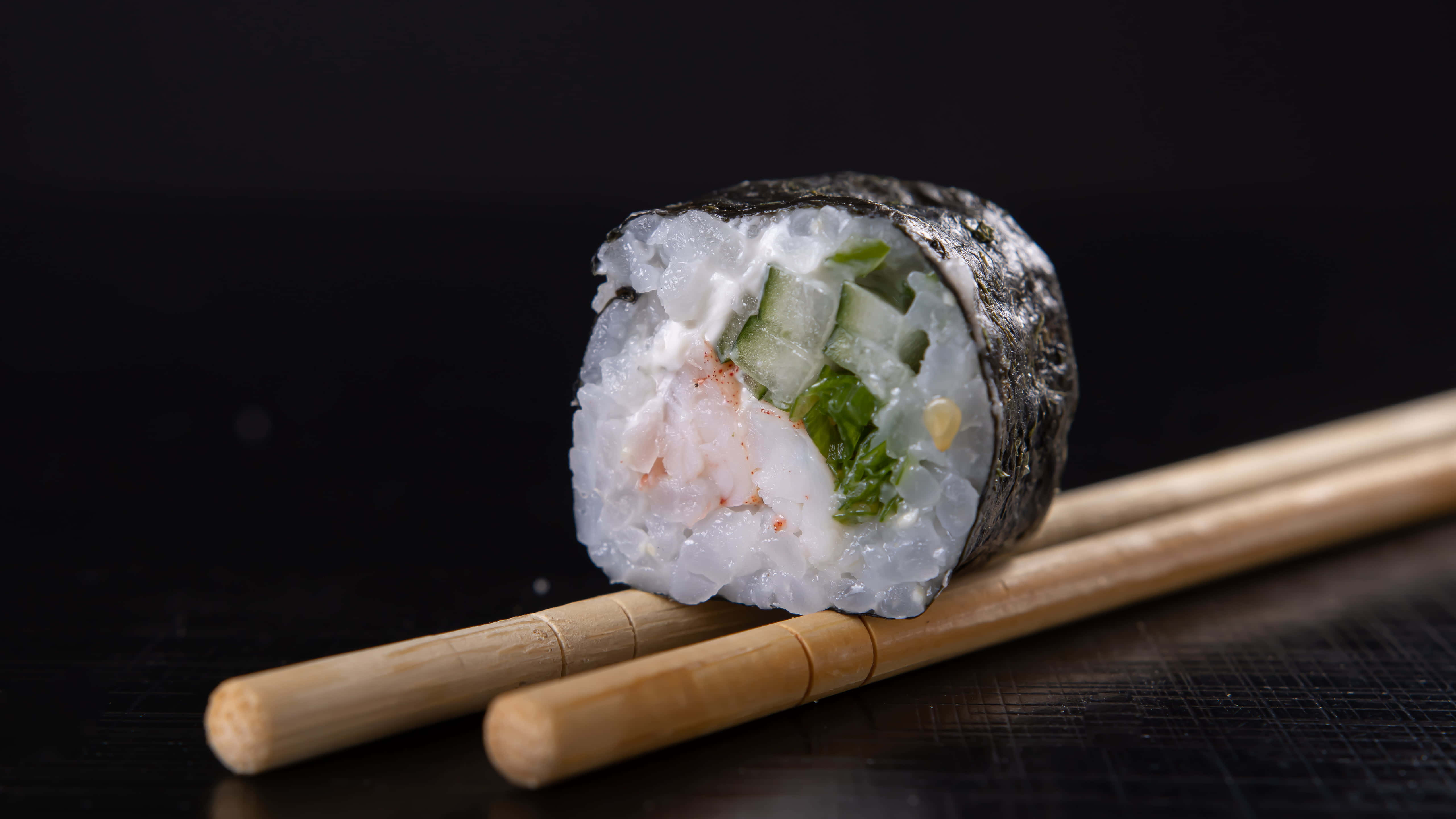 Types Of Sushi 桌布 壁紙 — 完美切好的壽司卷，內有蝦、小黃瓜和酪梨，放在木筷子上面，背景是深色有紋理的。