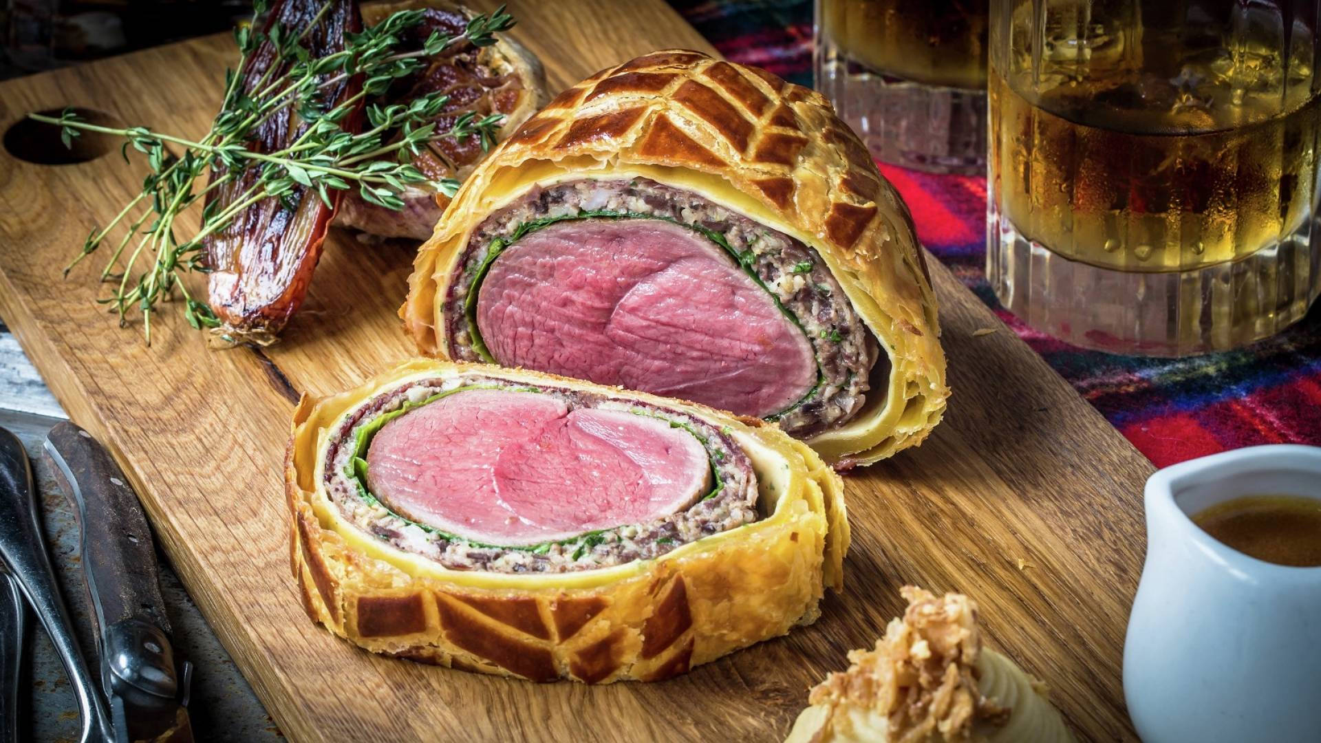 meat 桌布 壁紙 — 完美切片的威靈頓牛排，搭配金黃酥皮和新鮮百里香，放在質樸的木板上，旁邊有琥珀色液體的玻璃杯和醬汁船。