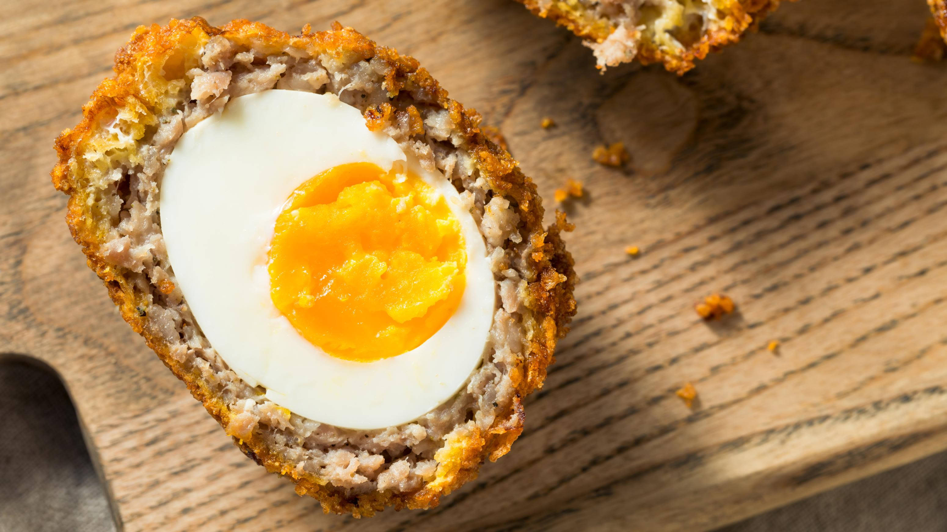 Egg 桌布 壁紙 — 完美切開的蘇格蘭蛋，在木板上展現出流心蛋黃，包裹在美味的肉餡和酥脆金黃的外皮中。