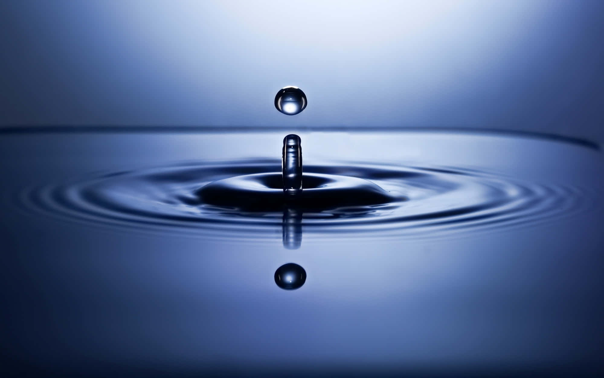 Water Drop 桌布 壁紙 — 完美捕捉的水滴懸浮在平靜的藍色水面上，形成同心漣漪和鏡面反射。