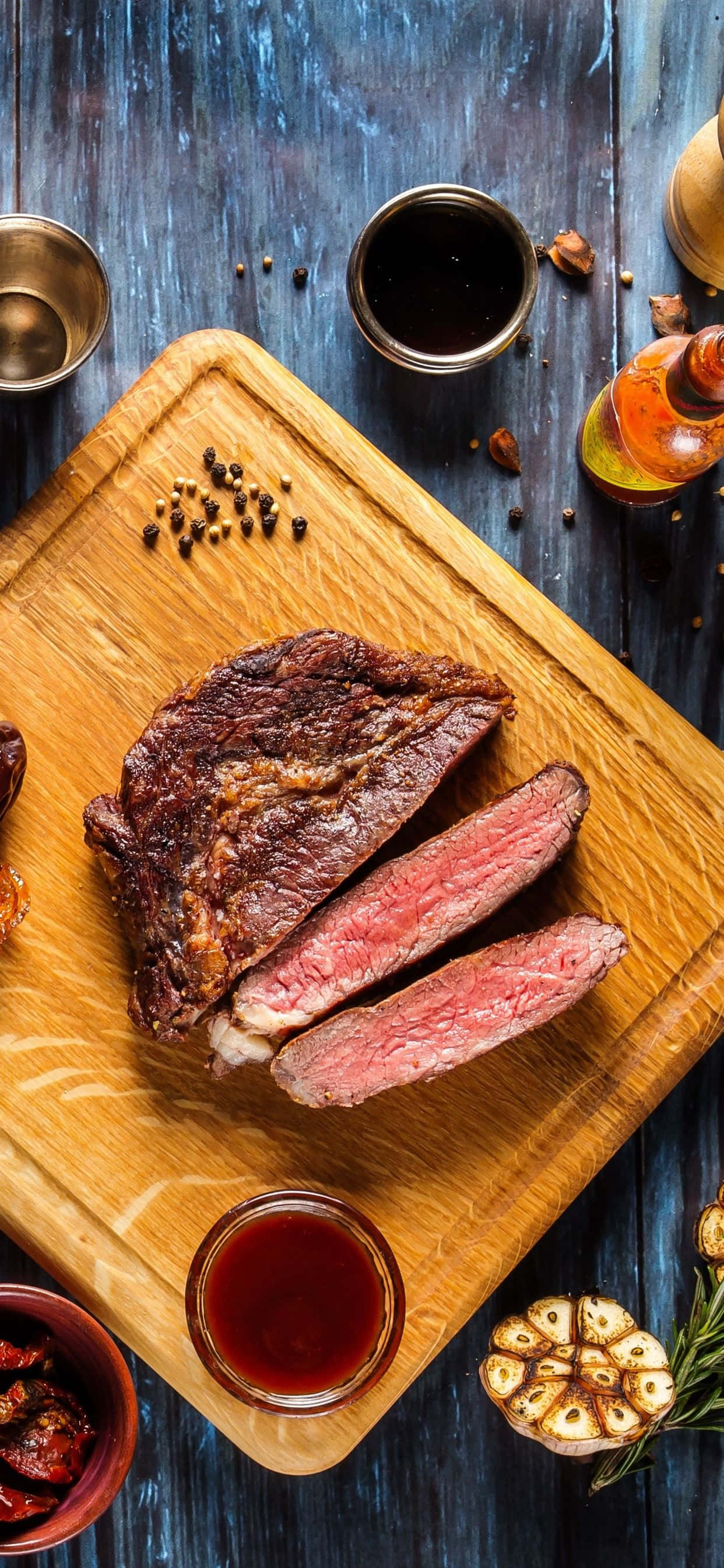 Cute Food iPhone 桌布 壁紙 — 完美煎烤的 medium-rare 牛排，切片放在木板上，配上大蒜、迷迭香和醬汁，置於鄉村風格的藍色桌子上。
