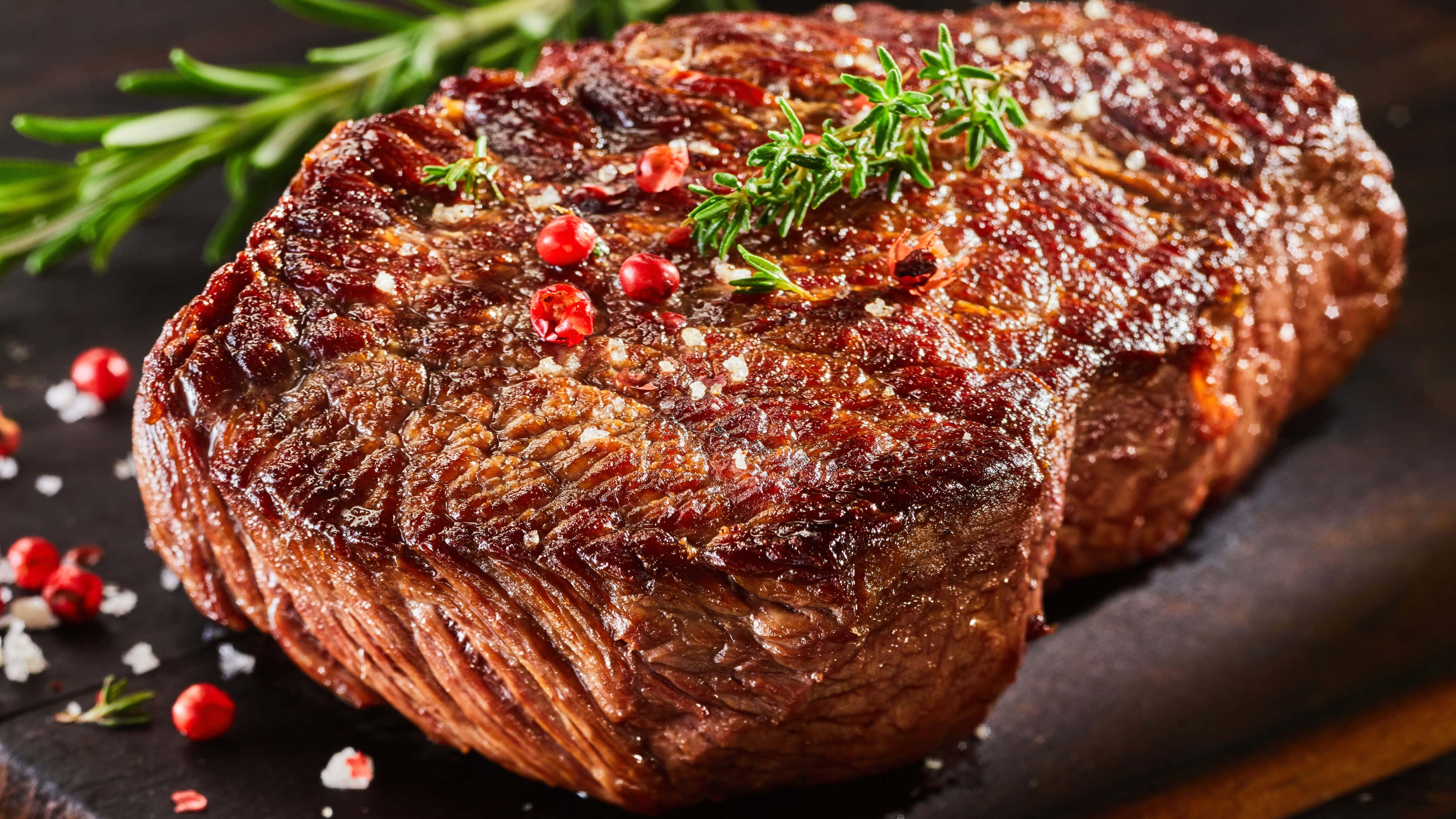 meat 桌布 壁紙 — 完美煎烤的牛排閃爍著鹽和胡椒的光澤，配上新鮮迷迭香和紅胡椒粒，放在深色木板上。