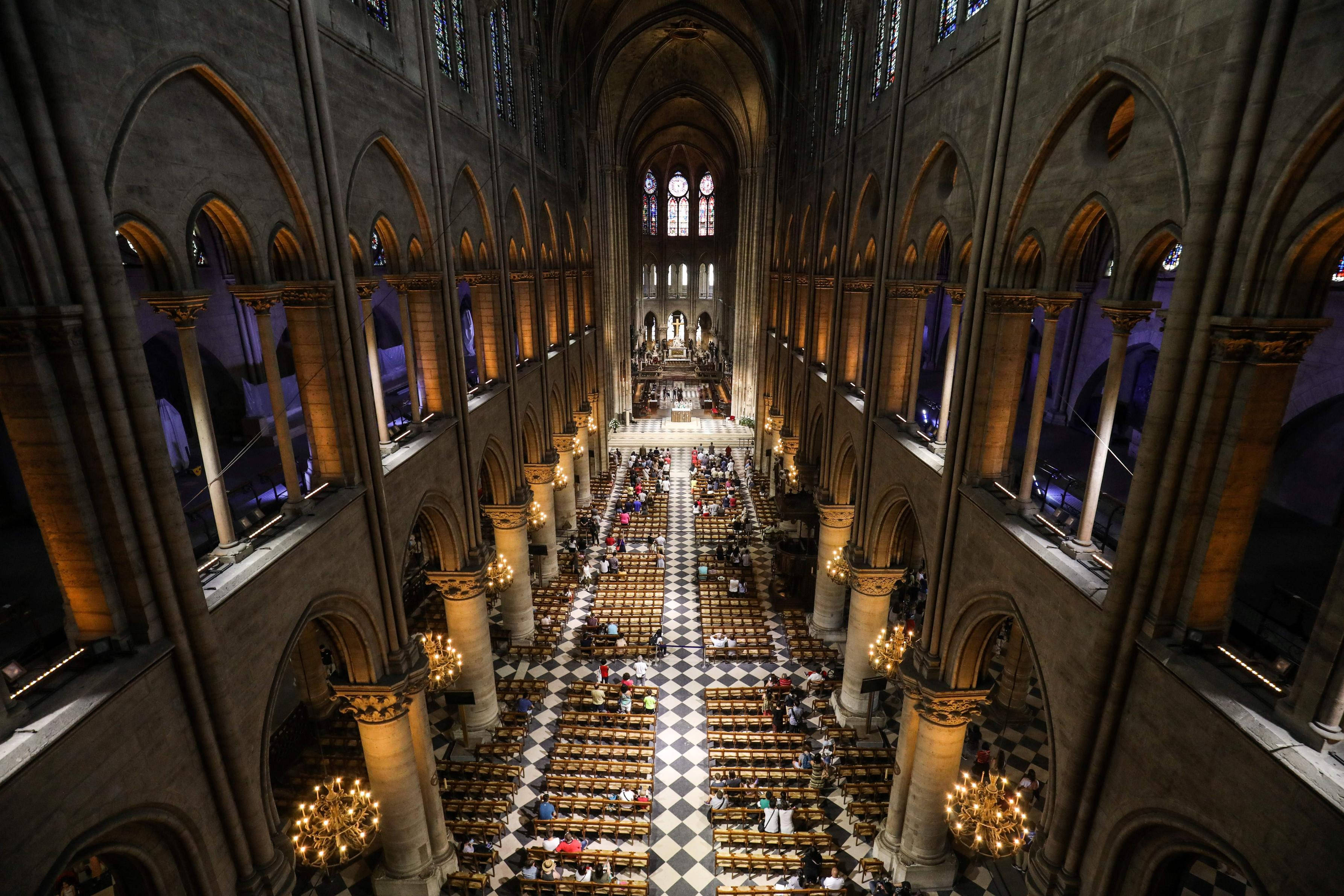 Notre Dame 桌布 壁紙 — 宏偉大教堂內部的鳥瞰圖，高聳的石拱門、棋盤格地板，以及信徒在溫暖金色燈光下坐在長椅上。