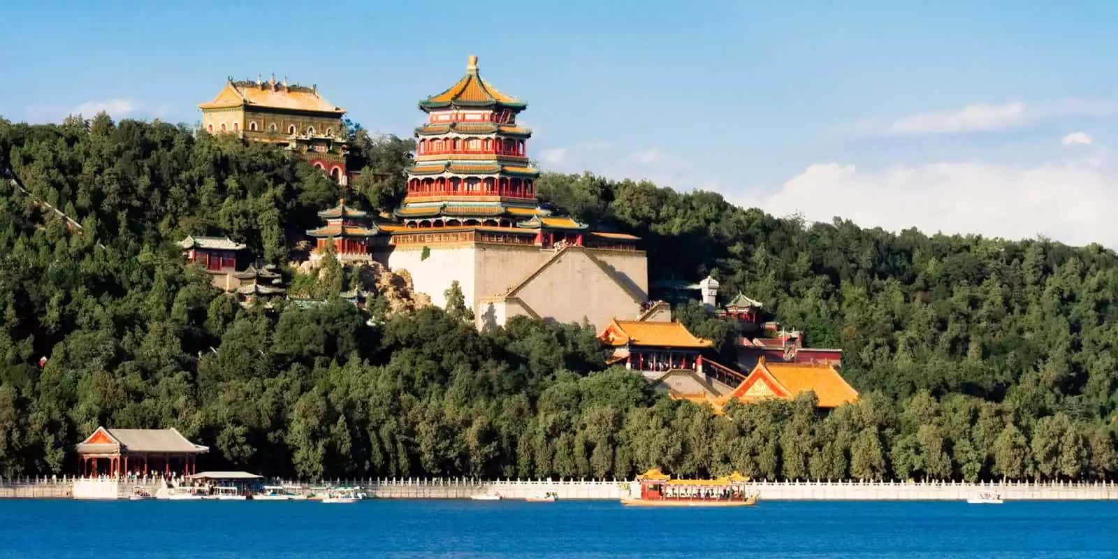 建築 桌布 壁紙 — 宏偉的傳統中國宮殿建築，金色屋頂矗立在鬱鬱蔥蔥的綠色山丘上，俯瞰著寧靜的藍色湖泊，湖上有船。