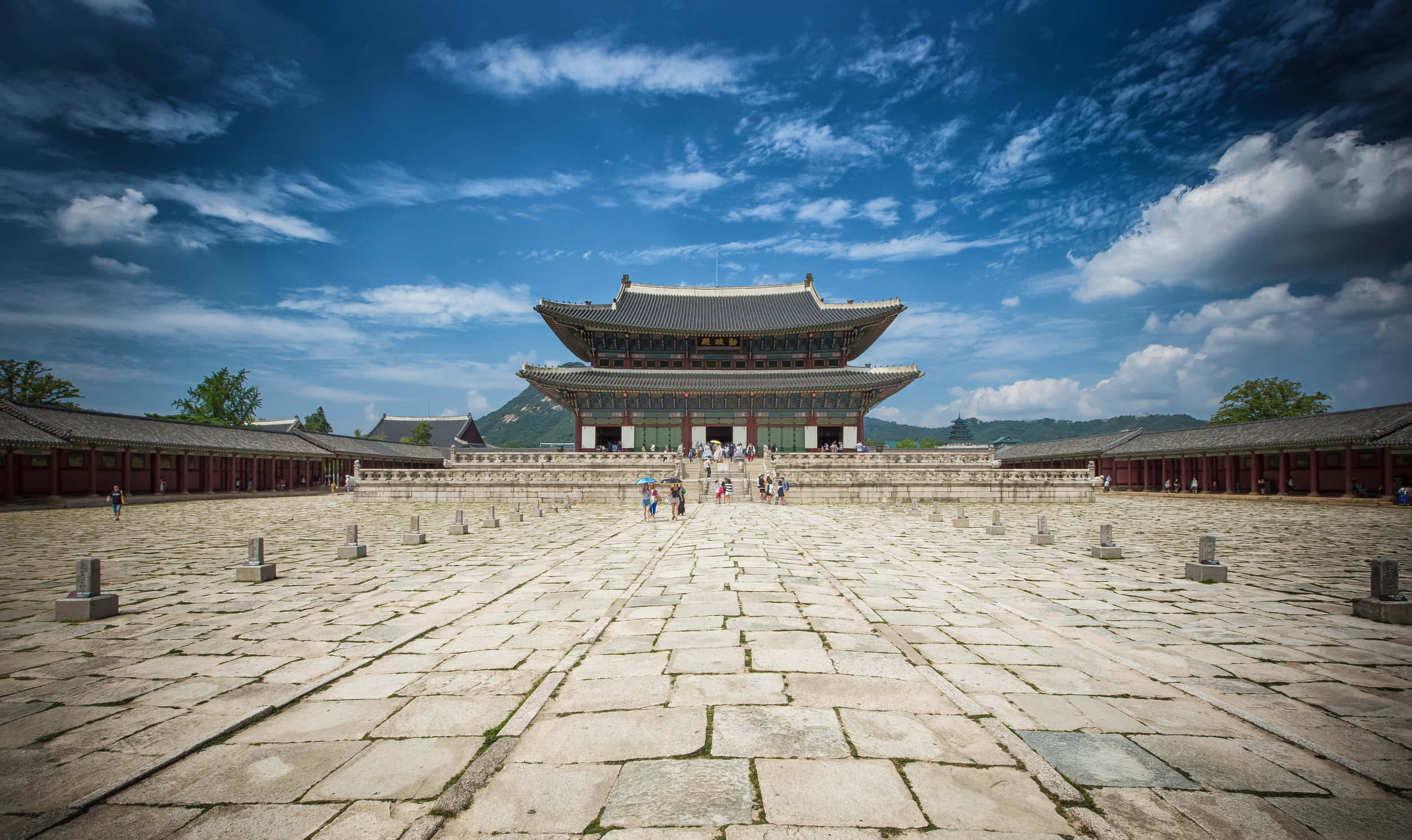 韓國 桌布 壁紙 — 宏偉的傳統韓國宮殿，擁有廣闊的石砌庭院，上方是湛藍的天空，點綴著幾朵白雲。