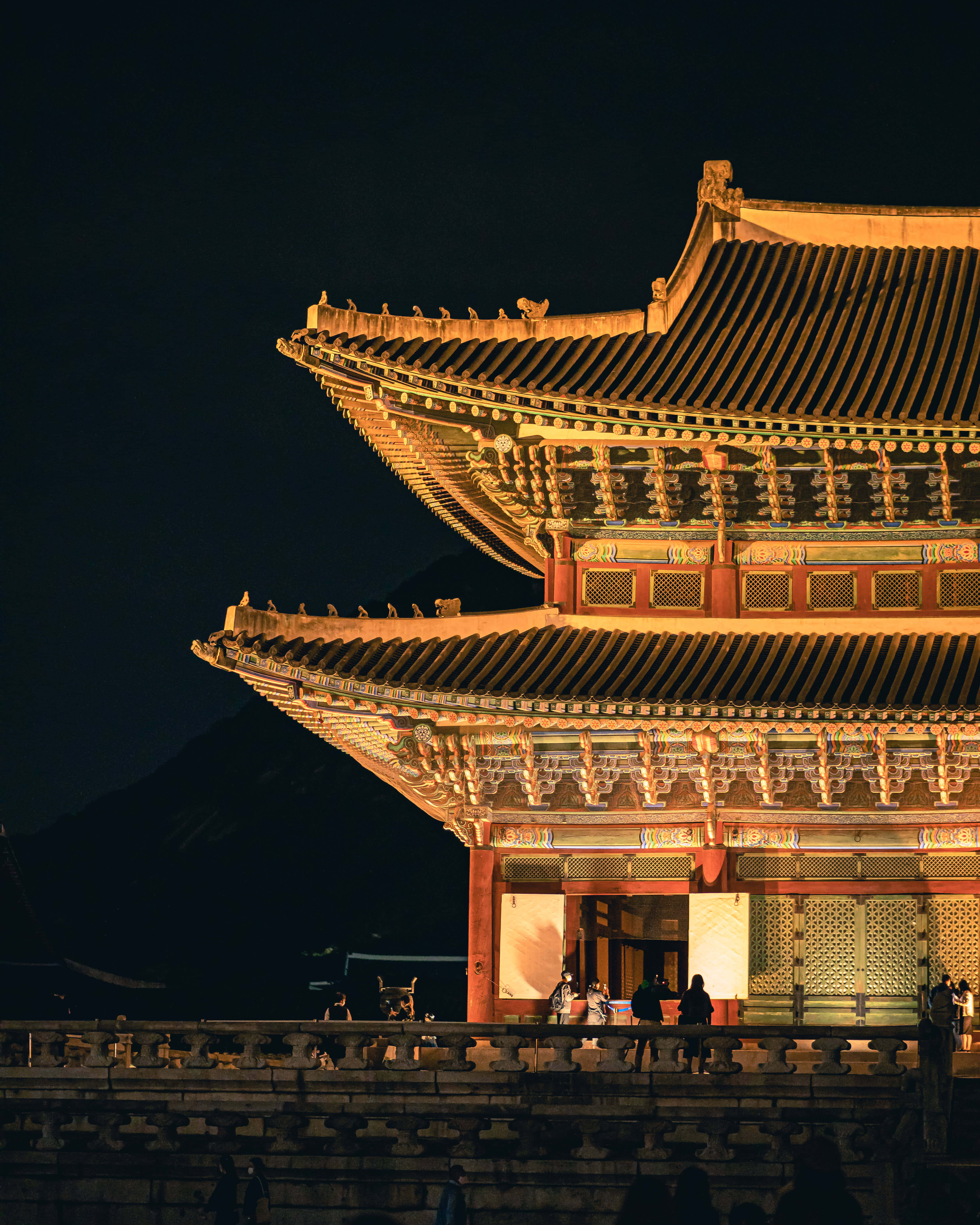 韓國 桌布 壁紙 — 宏偉的傳統韓國宮殿屋頂在夜燈下溫暖地發光，映襯著黑暗的天空，下方有剪影般的遊客。