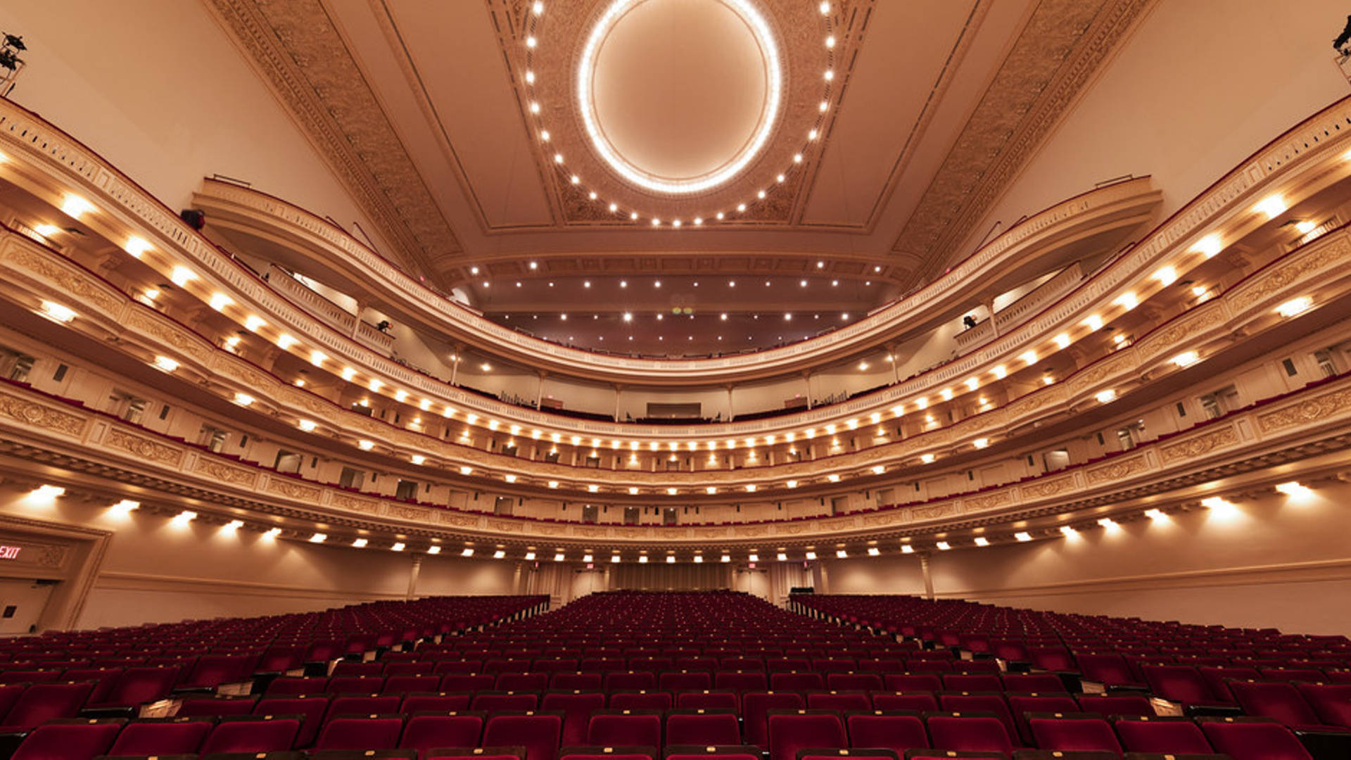 theatre 桌布 壁紙 — 宏偉的劇院內部，設有分層陽台、華麗天花板和沐浴在溫暖金色光線中的紅色座位排。