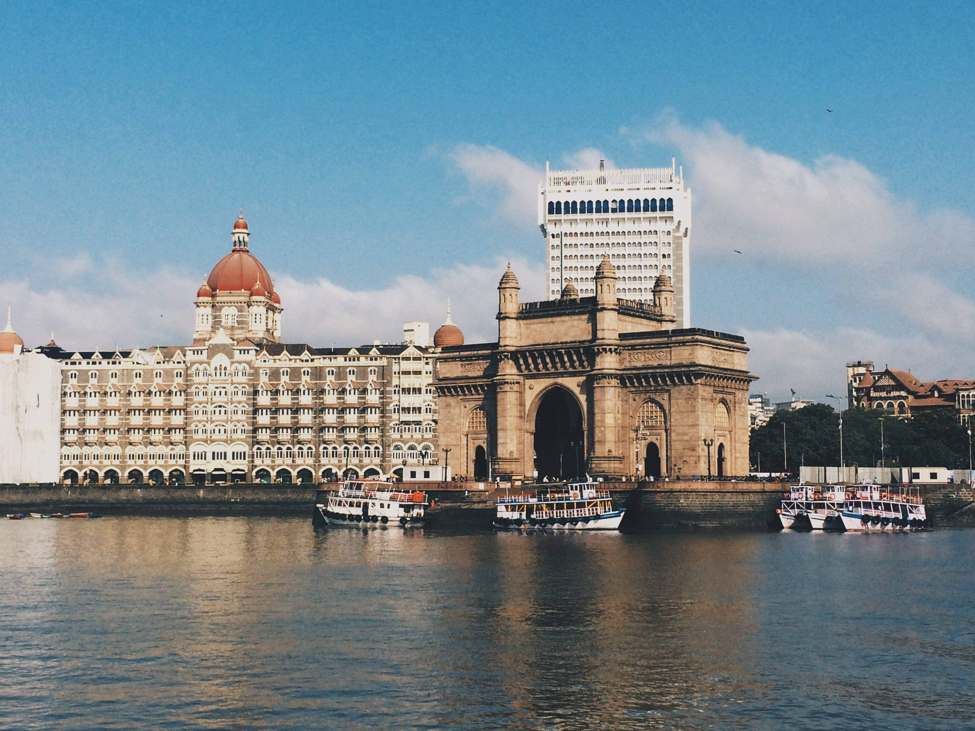 Mumbai 桌布 壁紙 — 宏偉的印度門拱門矗立在水邊，泰姬瑪哈皇宮酒店和船隻在明亮的藍天下。