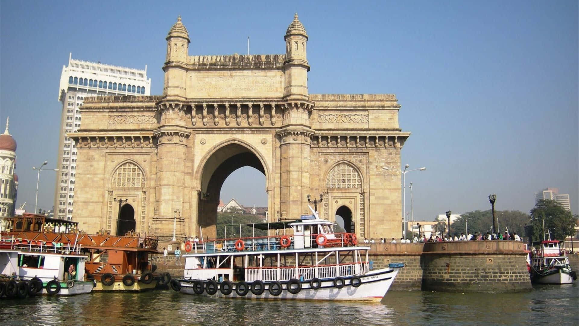 Mumbai 桌布 壁紙 — 宏偉的印度門拱門矗立在繁忙的港口之上，港內有船隻和現代建築，天空清澈湛藍。