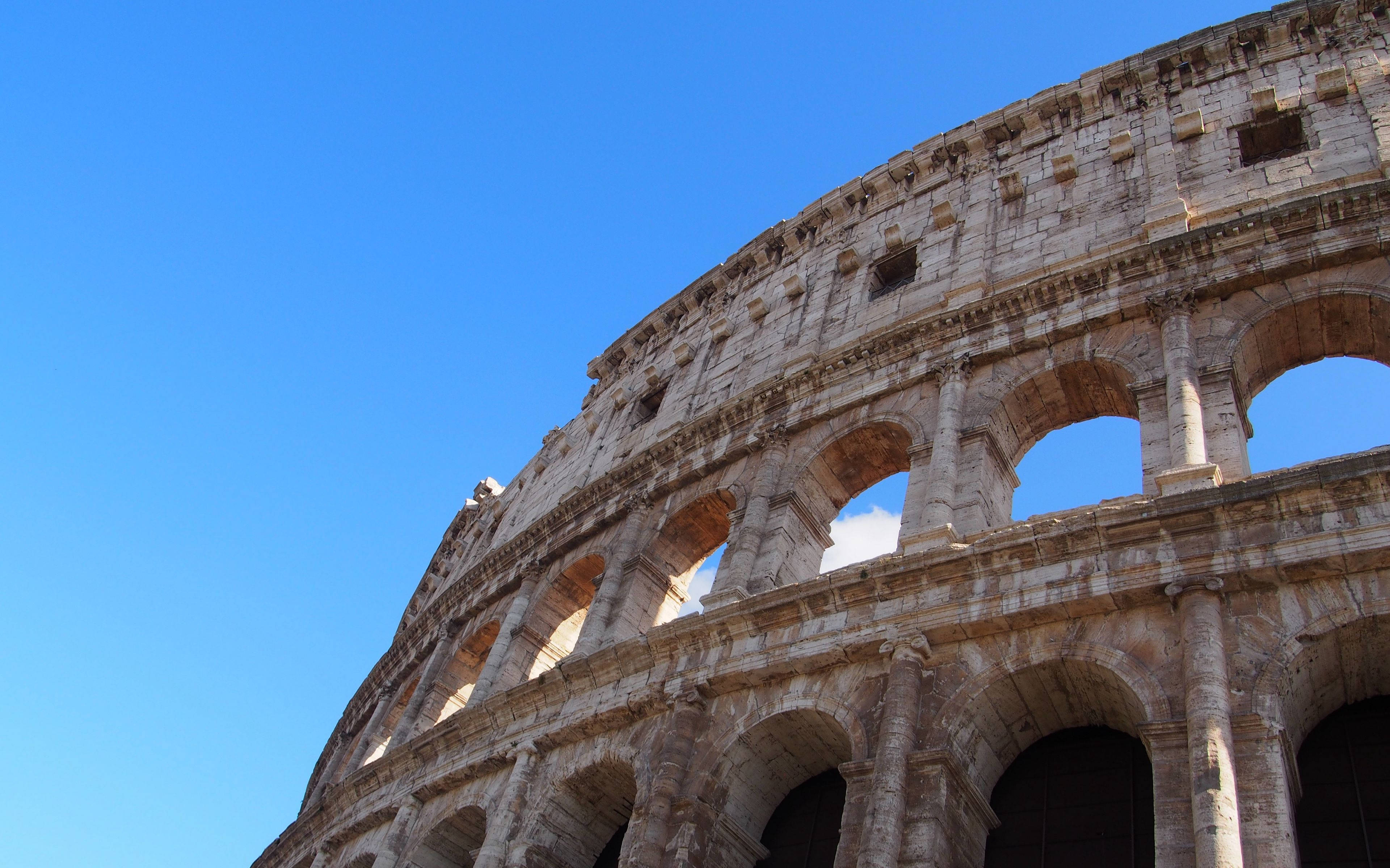 Rome 桌布 壁紙 — 宏偉的古羅馬競技場在晴朗的藍天下雄偉地聳立，石拱門沐浴在陽光中。