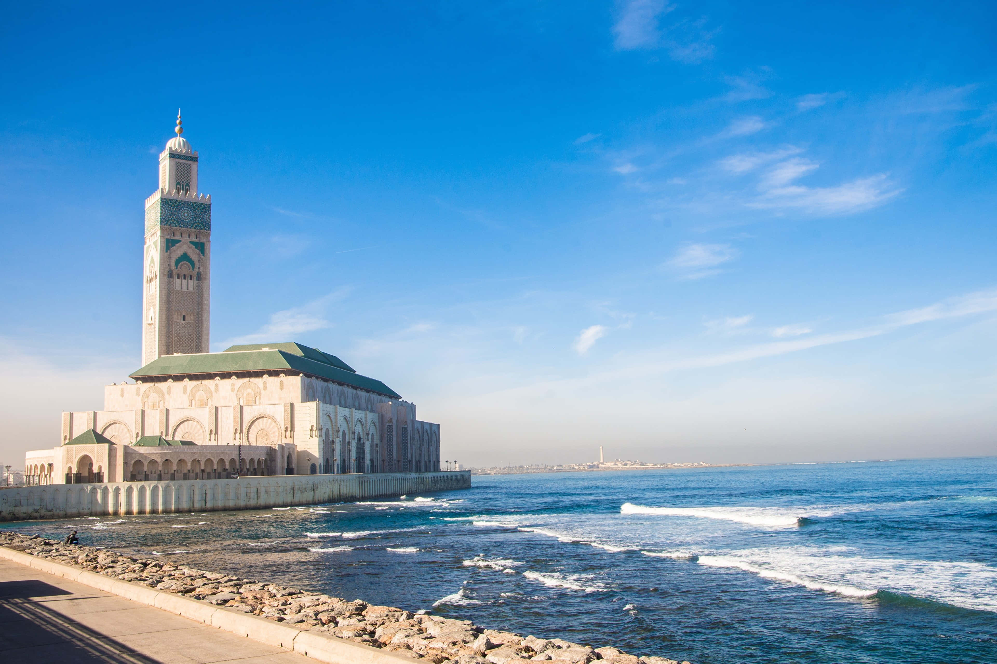 Mosque 桌布 壁紙 — 宏偉的哈桑二世清真寺在清澈的藍天下雄偉地矗立在海邊，溫柔的海浪在附近拍打。
