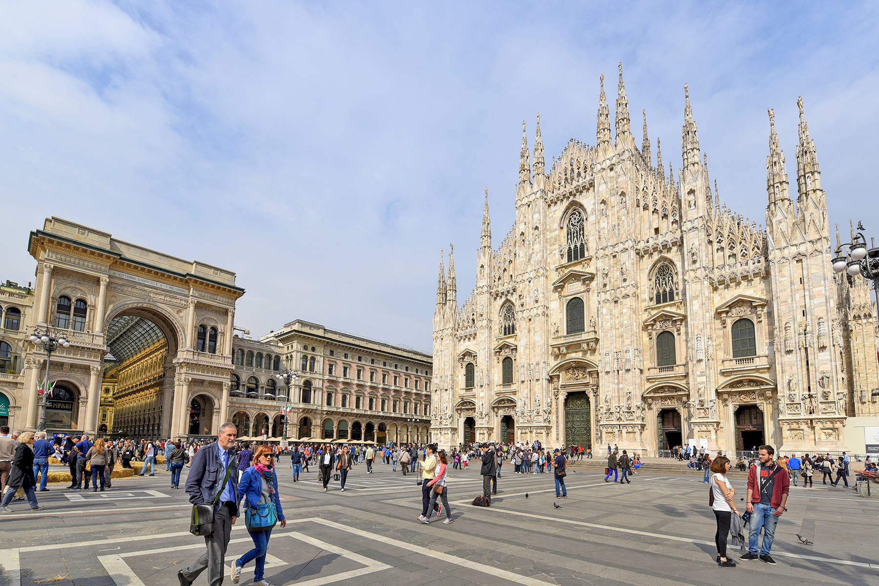 Milan 桌布 壁紙 — 宏偉的哥德式大教堂，尖塔和華麗的外牆，在部分多雲的天空下，主宰著一個擠滿遊客的繁忙廣場。