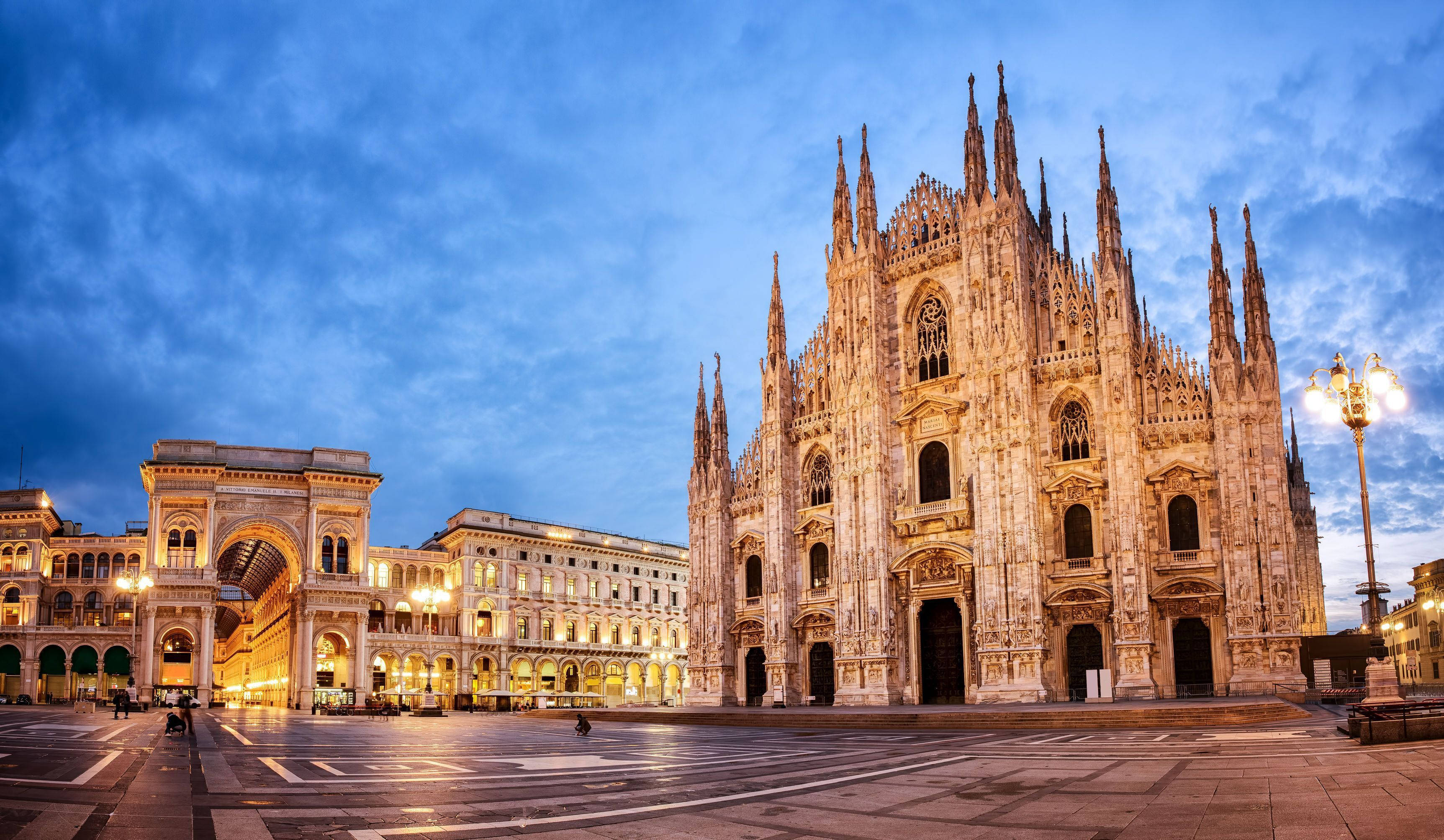 教堂 桌布 壁紙 — 宏偉的哥德式大教堂在黃昏時分被溫暖的燈光照亮，襯著深藍色的天空和空曠的廣場。