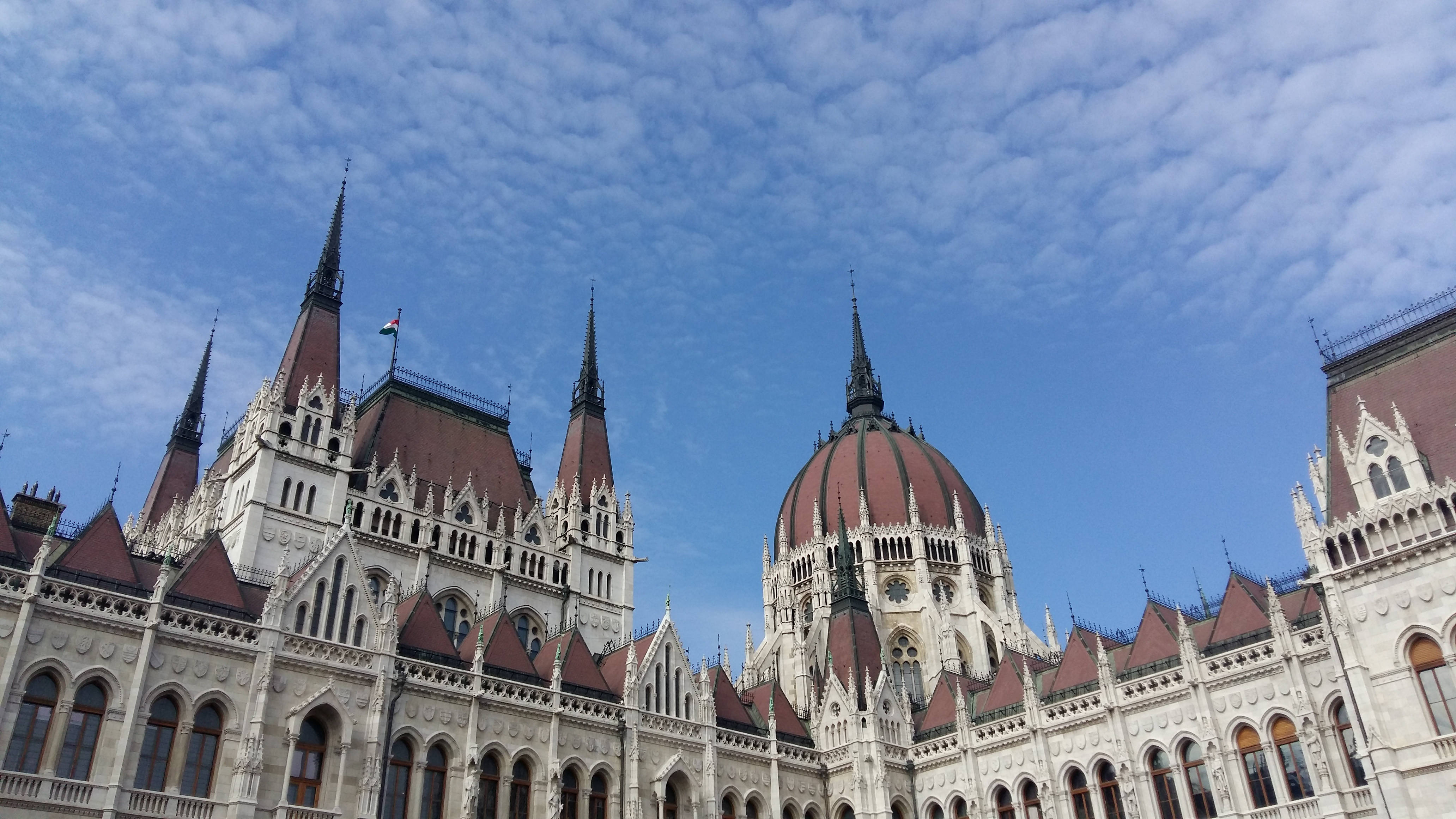 Hungary 桌布 壁紙 — 宏偉的哥德式議會大樓，擁有紅色瓦片屋頂和尖塔，背景是明亮的藍天和散落的雲朵。