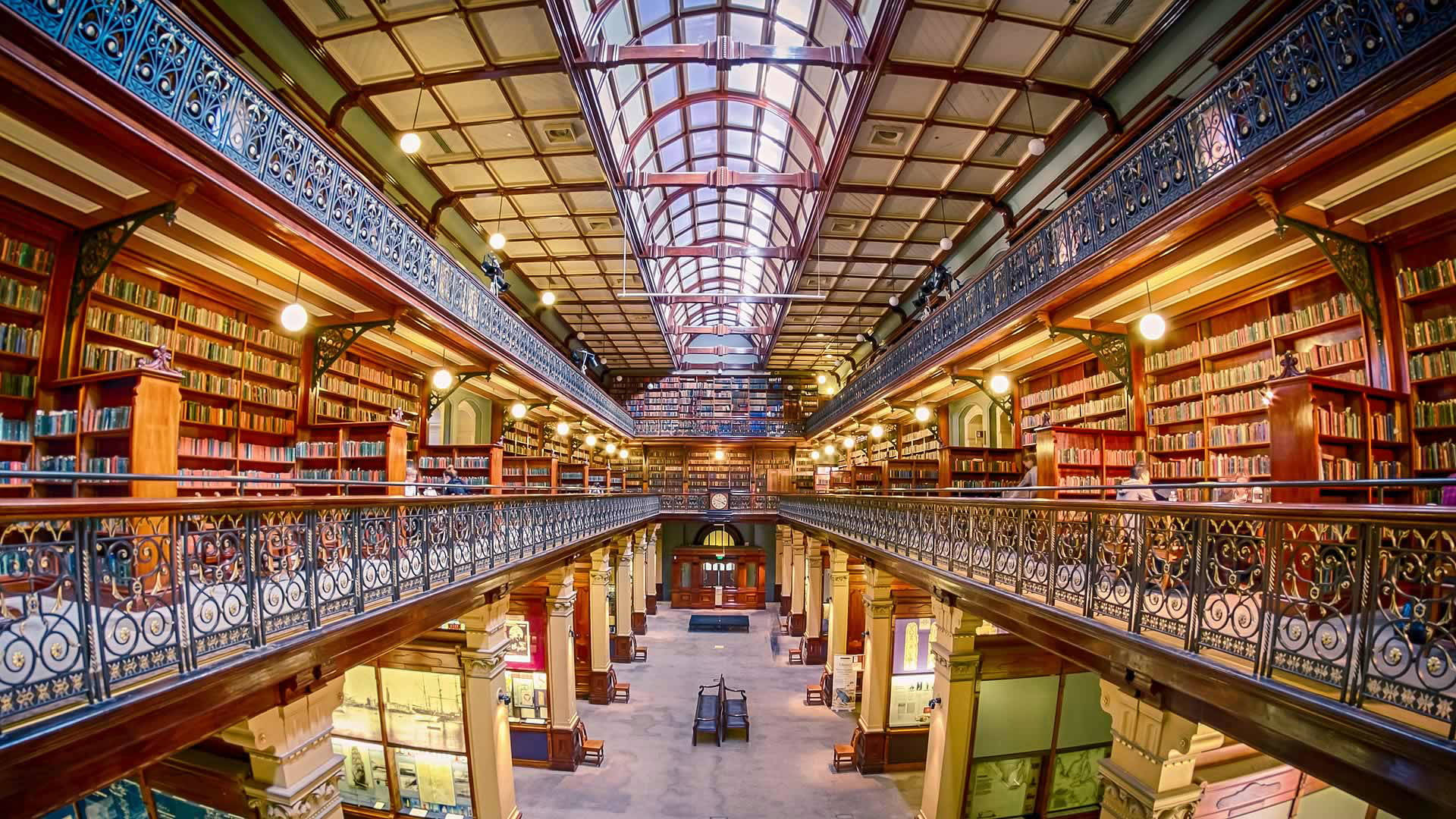Adelaide 桌布 壁紙 — 宏偉的圖書館，擁有高聳的書架、華麗的鐵欄杆和玻璃穹頂天花板，溫暖的光線灑滿整個空間。