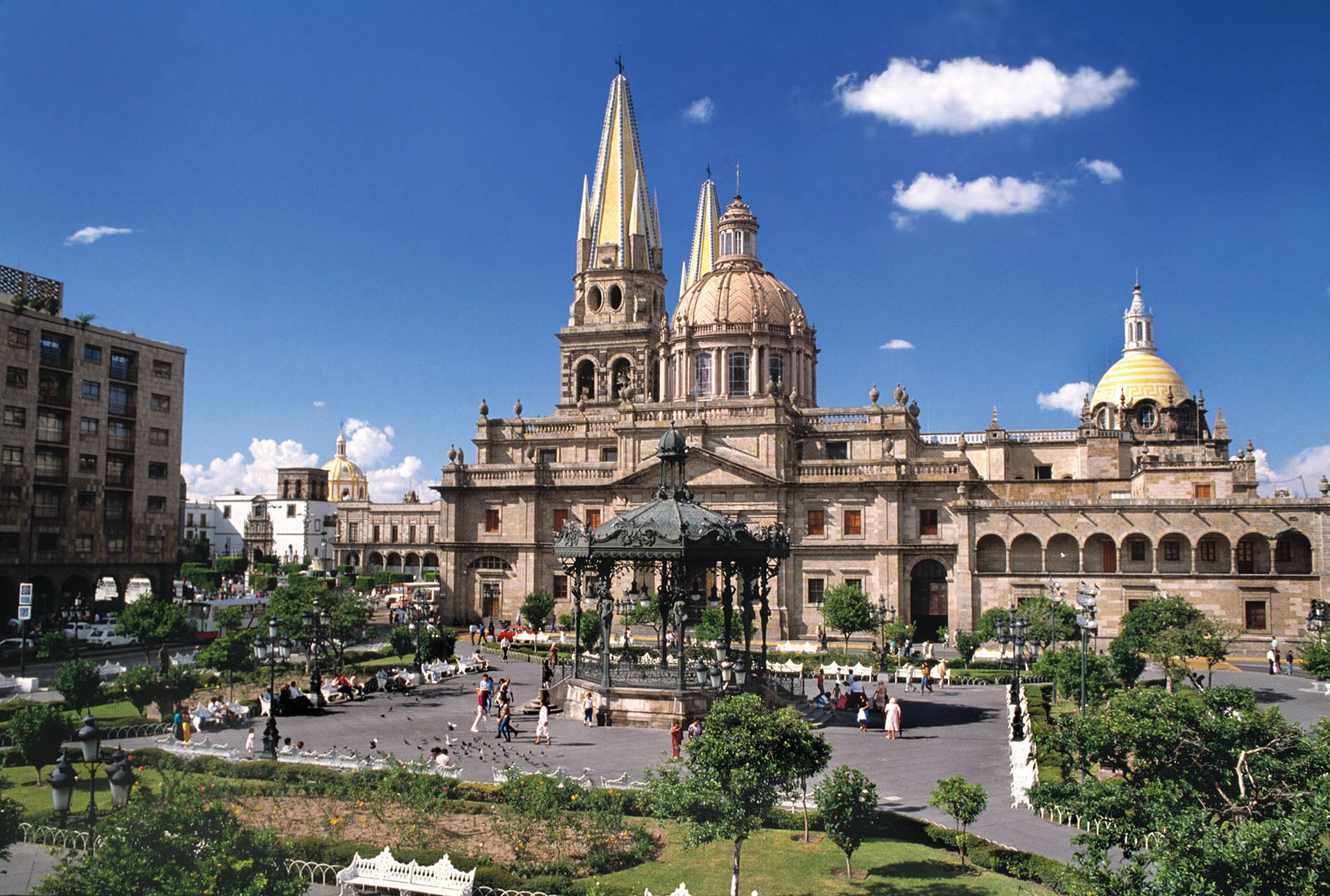 Guadalajara 桌布 壁紙 — 宏偉的大教堂，金色圓頂和尖塔聳立在陽光明媚的廣場上，廣場上滿是人們和華麗的綠植，背景是明亮的藍天。