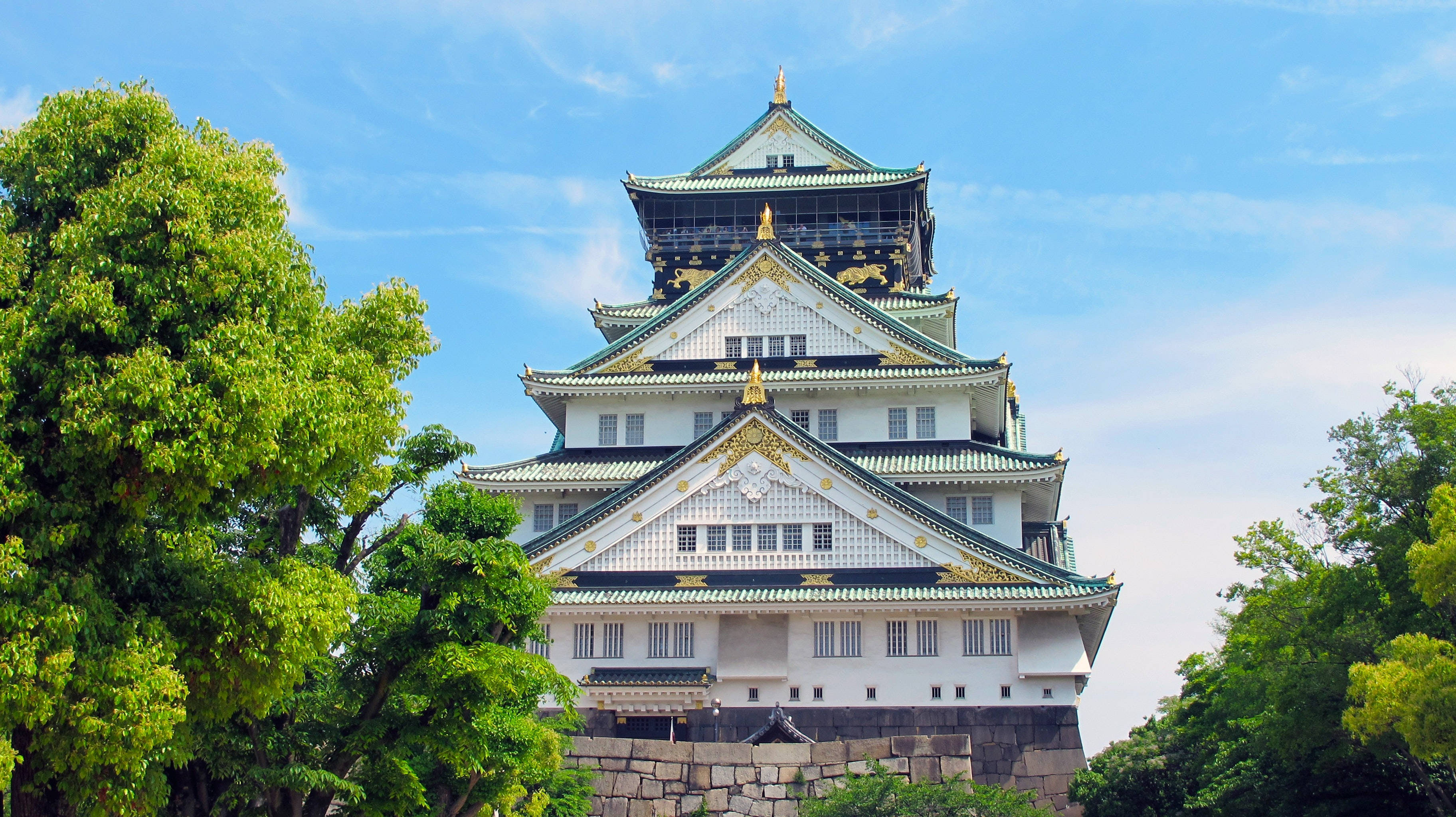 Osaka Castle 桌布 壁紙 — 宏偉的大阪城在晴朗的日子裡，在藍天白雲和茂密綠樹的映襯下巍然聳立。