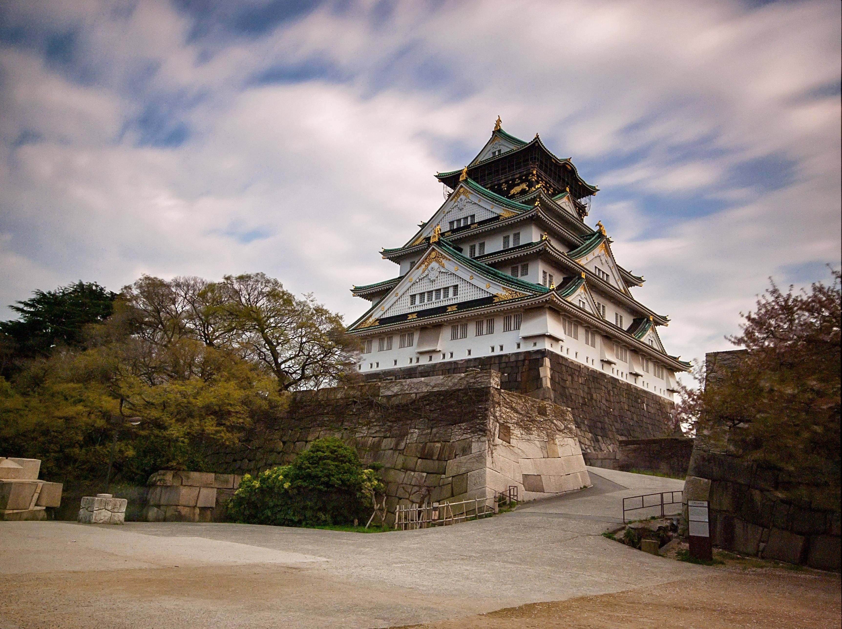 Osaka Castle 桌布 壁紙 — 宏偉的大阪城在陰雲密布的天空下巍然聳立，秋樹環繞著它的石基和傳統瓦頂。