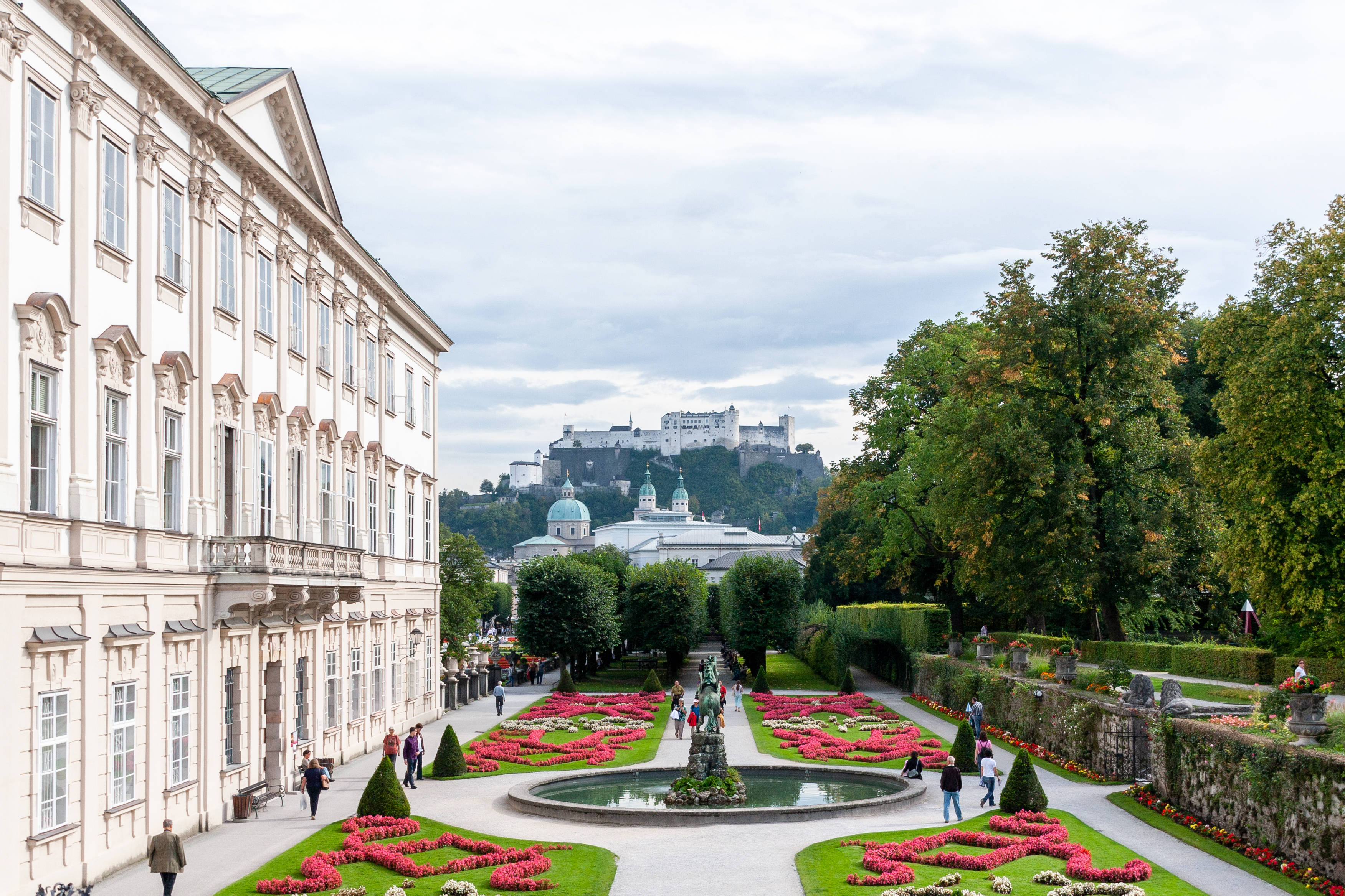 Austria 桌布 壁紙 — 宏偉的宮殿花園，對稱的花壇和中央噴泉，通向山丘上的城堡，天空多雲。