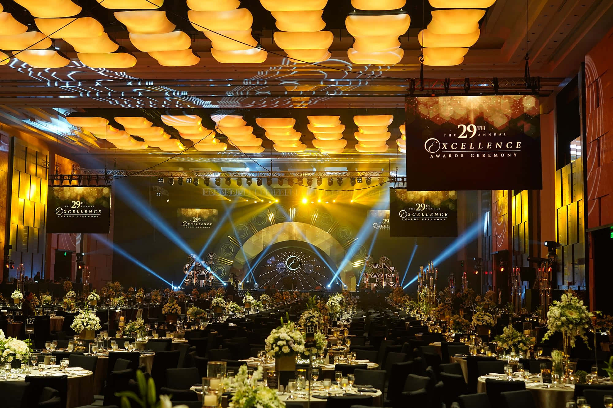 Corporate Event 桌布 壁紙 — 宏偉的宴會廳內，在溫暖的吊燈和充滿活力的舞台光束下，整齊擺放著優雅的餐桌，為第29屆卓越獎頒獎典禮而設。