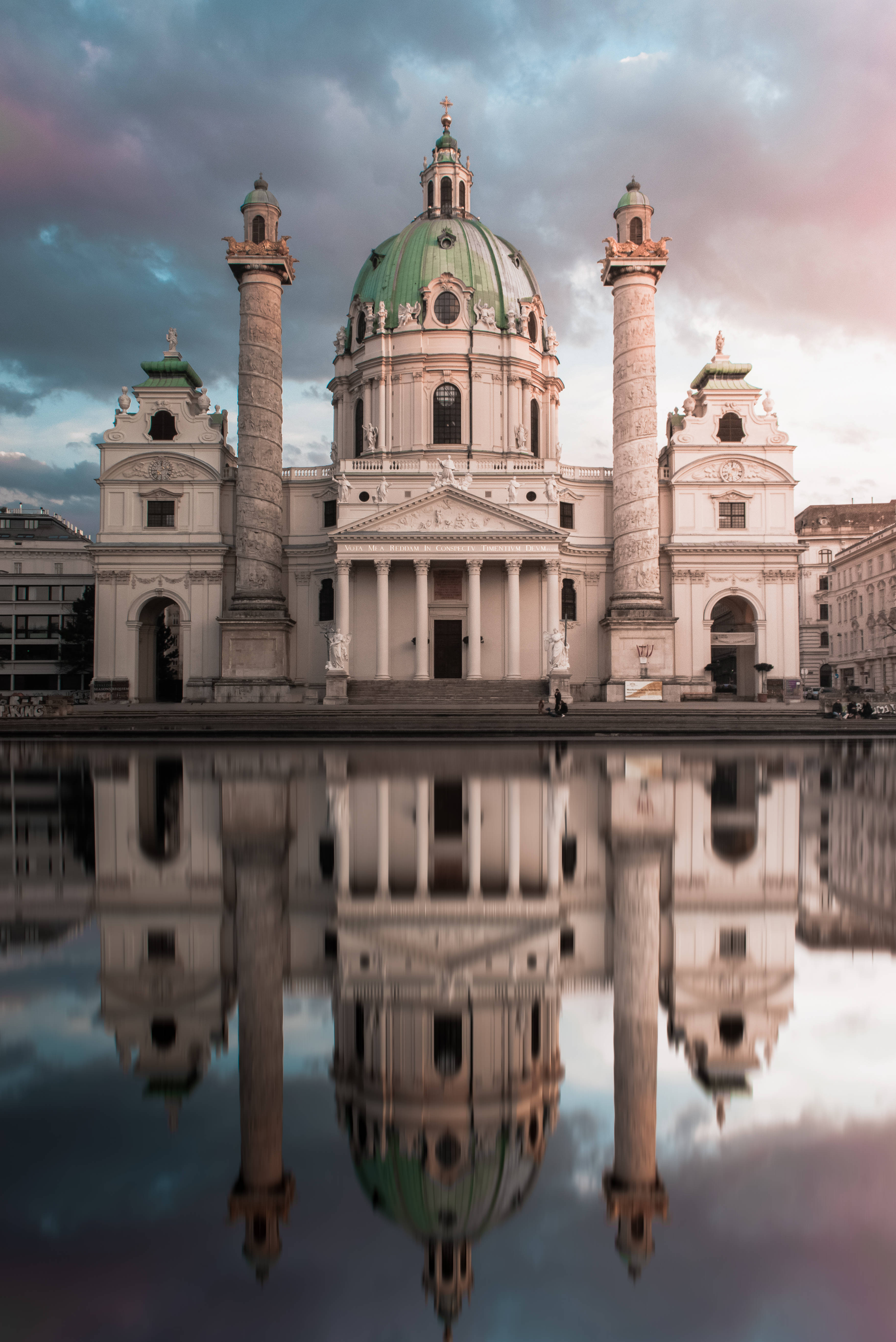 Austria 桌布 壁紙 — 宏偉的巴洛克式大教堂，擁有綠色圓頂和兩側柱廊，在戲劇性的暮色天空下完美倒映在平靜的水面上。