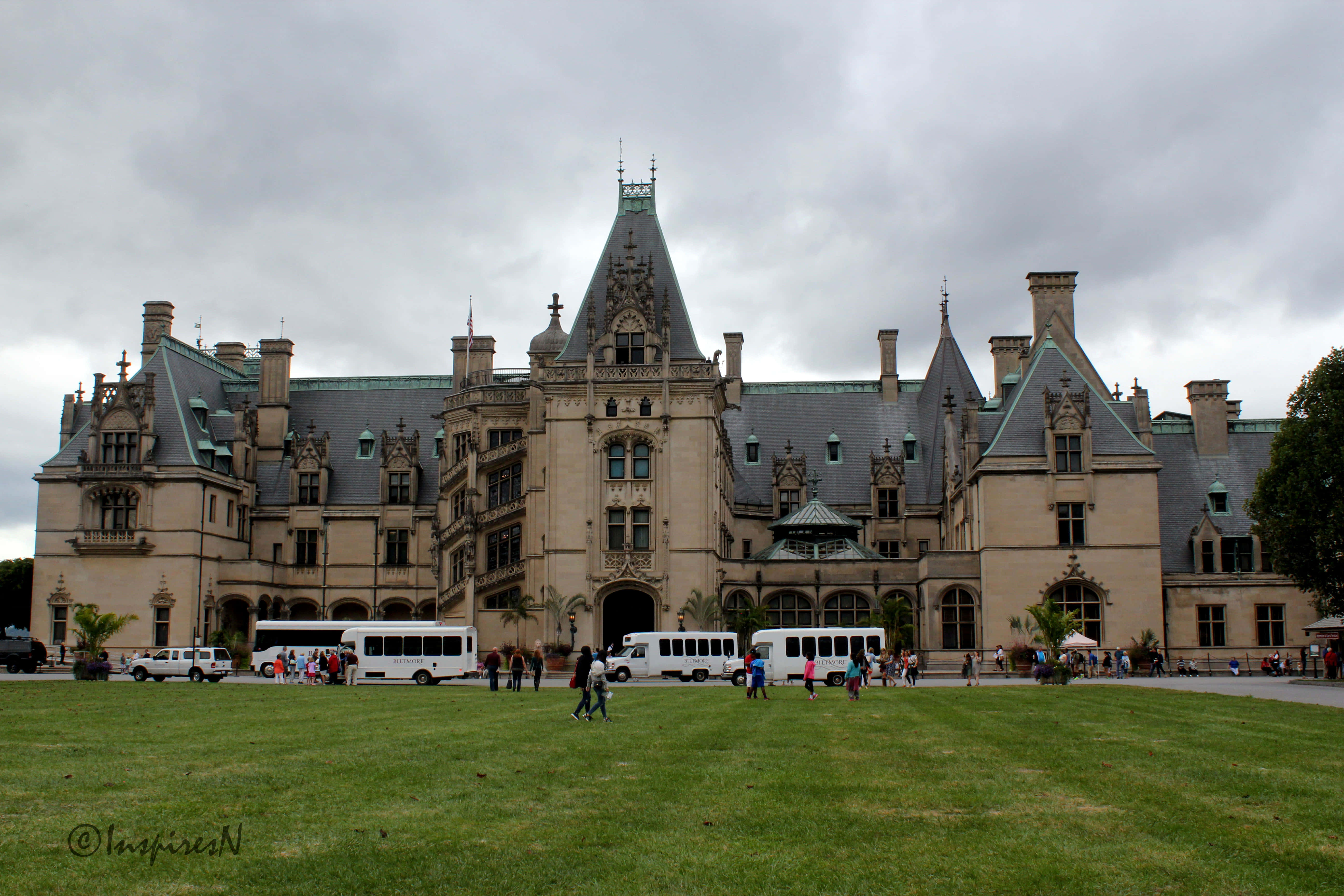 Biltmore Estate 桌布 壁紙 — 宏偉的布雜藝術風格大宅，帶有華麗的角樓和灰色石板屋頂，陰天，修剪整齊的草坪上有遊客和旅遊巴士。