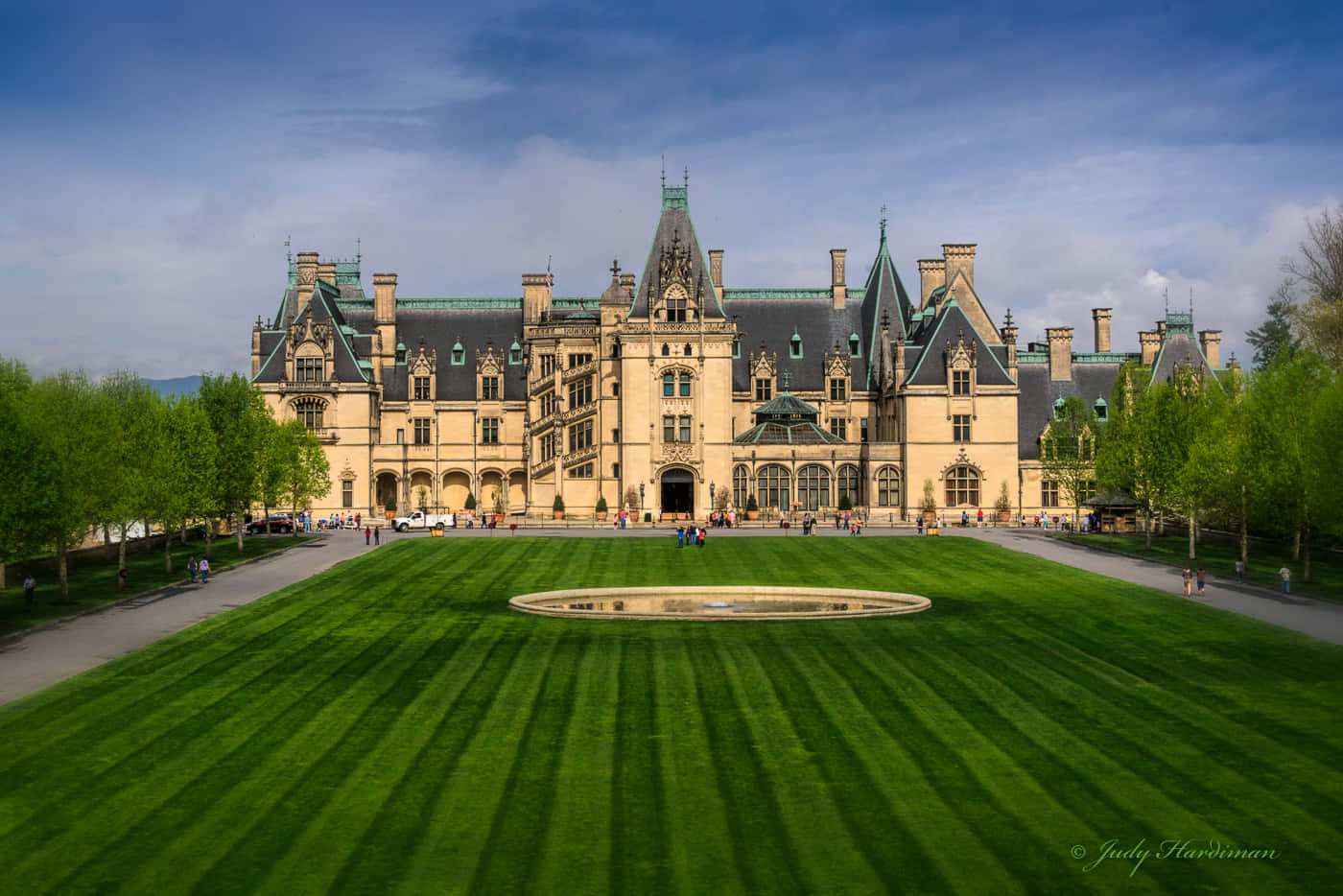 Biltmore Estate 桌布 壁紙 — 宏偉的布雜藝術風格大宅，擁有綠色屋頂塔樓，雄偉地矗立在廣闊的條紋草坪和中央噴泉後方，天空部分多雲。