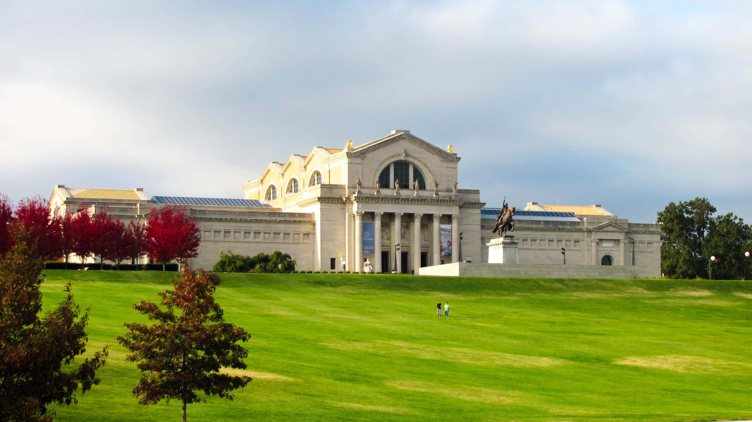 Museum 桌布 壁紙 — 宏偉的新古典主義博物館，擁有柱子和雕像，矗立在廣闊的綠色草坪上，天空多雲，附近有紅葉樹木。