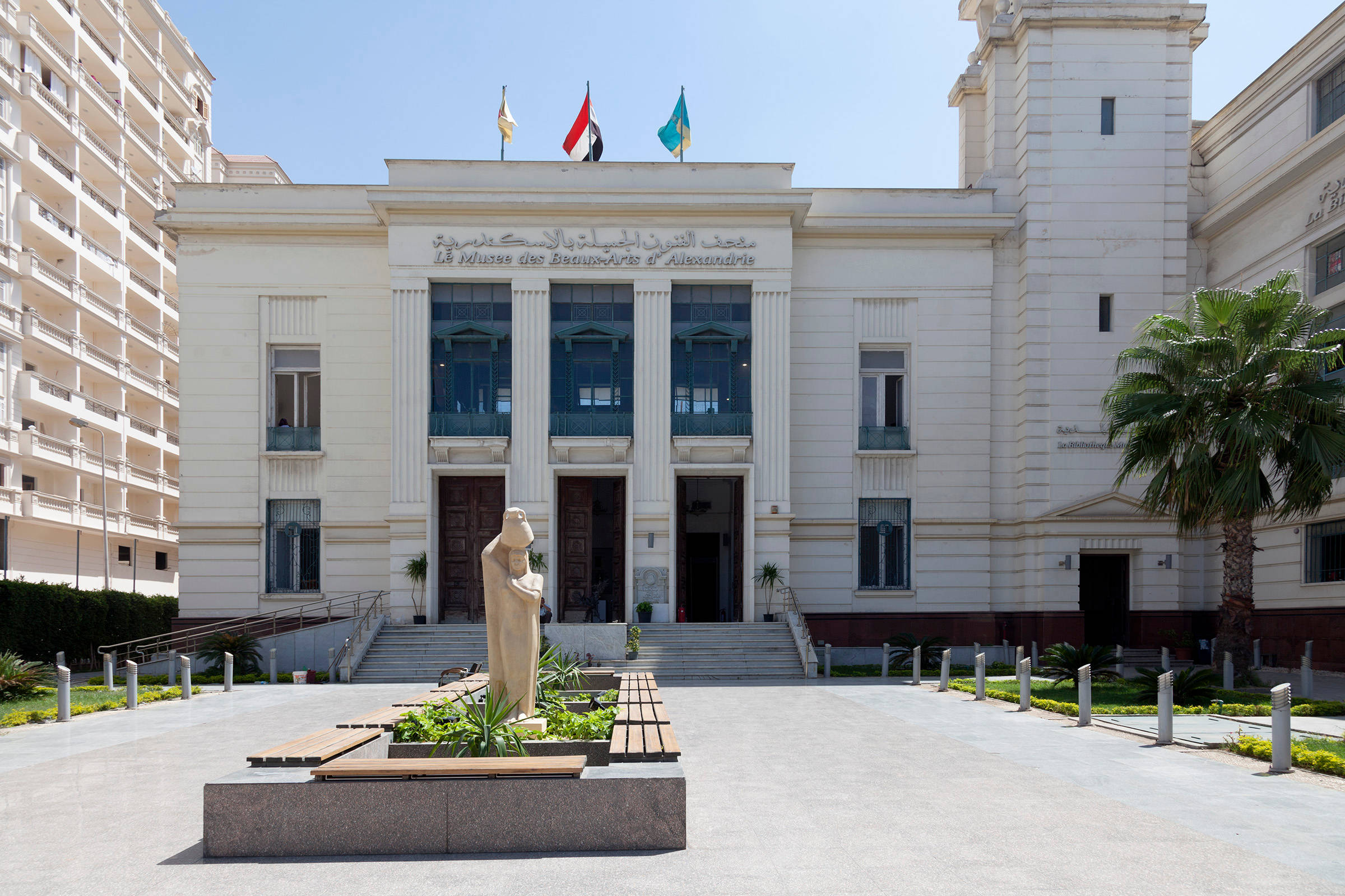 Alexandria 桌布 壁紙 — 宏偉的新古典主義博物館外牆，柱子和旗幟在燦爛陽光下，兩旁有棕櫚樹和中央雕像。