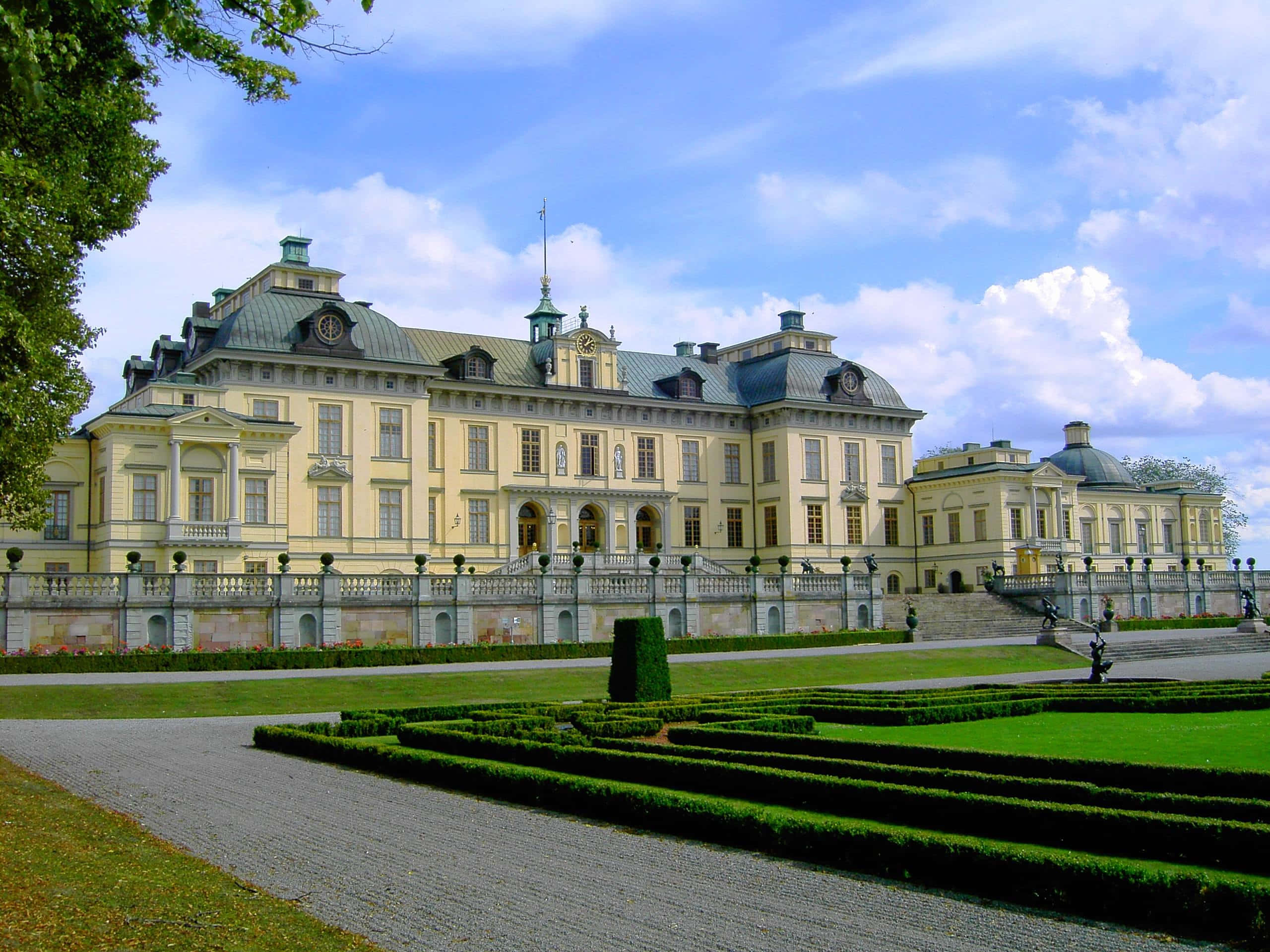 Drottningholm Palace 桌布 壁紙 — 宏偉的新古典主義宮殿，擁有對稱的翼樓和綠色圓頂，矗立在藍天白雲下，周圍是修剪整齊的花園和石徑。