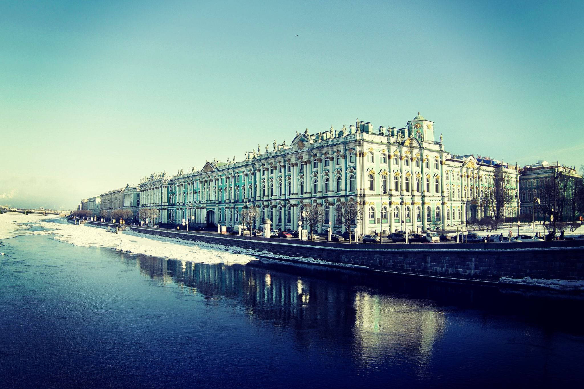 Saint Petersburg 桌布 壁紙 — 宏偉的新古典主義宮殿，金色點綴，在清澈的藍色冬日天空下倒映在冰冷的河流中。