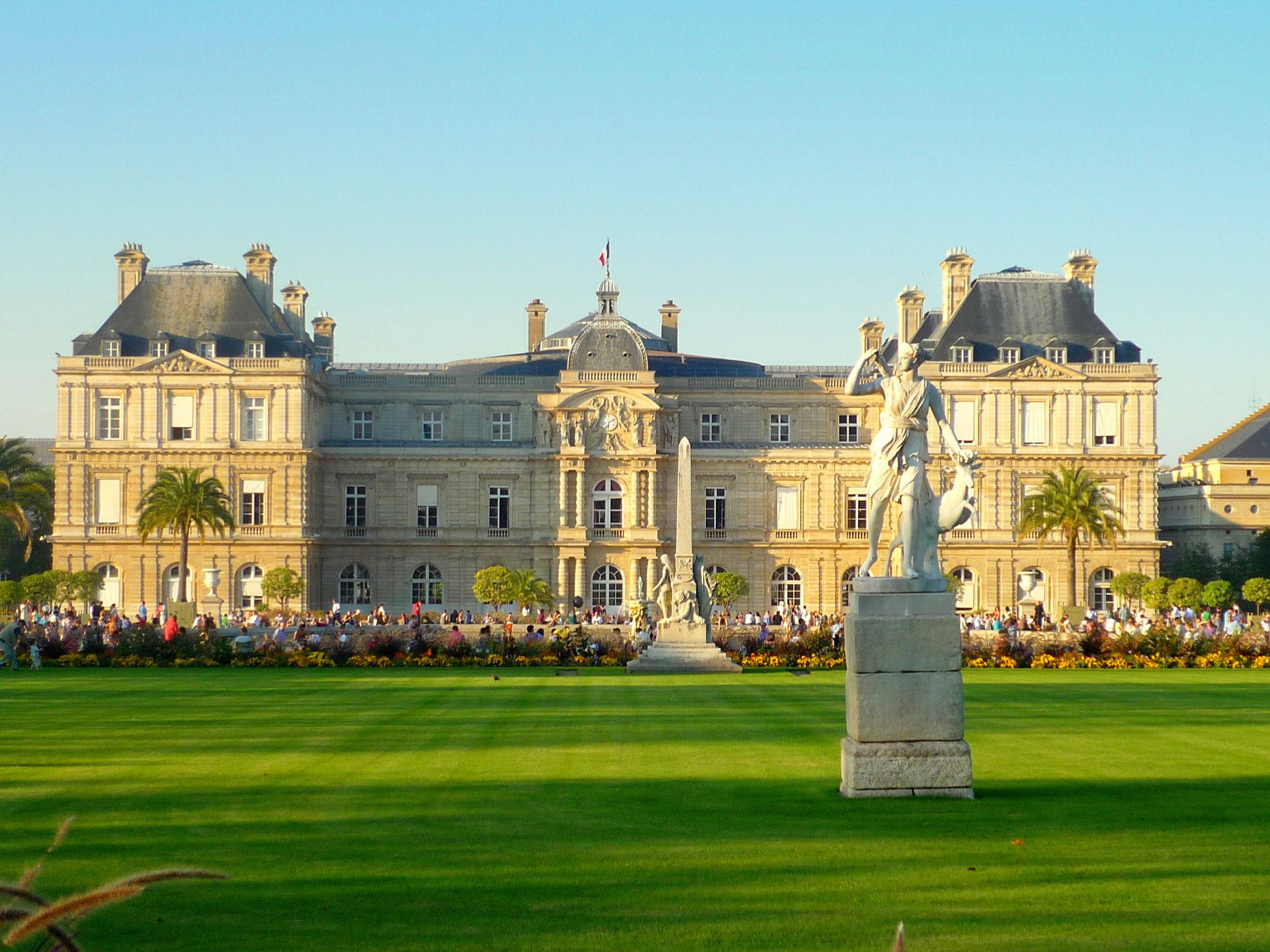 Luxembourg 桌布 壁紙 — 宏偉的新古典主義宮殿沐浴在金色的日落光芒中，前景是茂密的綠色草坪和雕像。