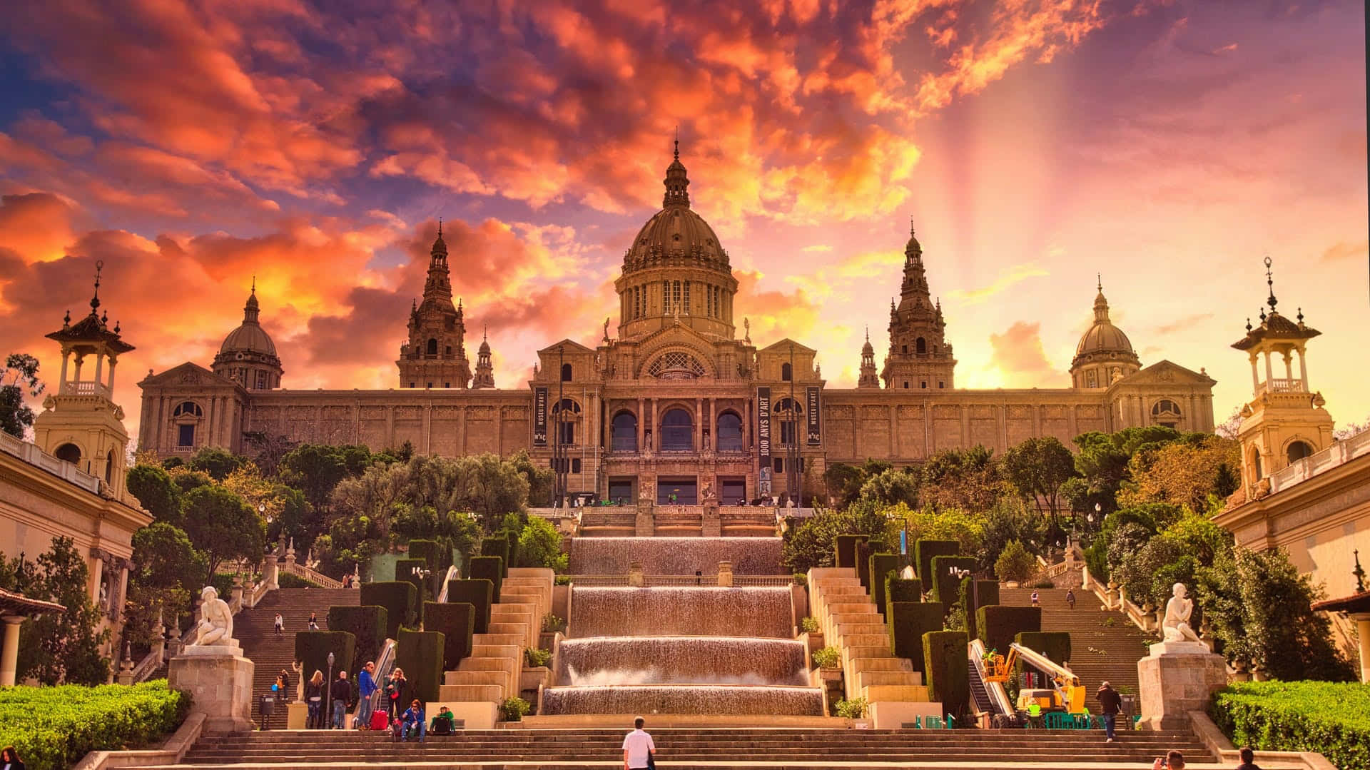Barcelona 桌布 壁紙 — 宏偉的新古典主義建築，圓頂和塔樓沐浴在熾熱的日落色調中，下方是層疊的噴泉和修剪整齊的花園。
