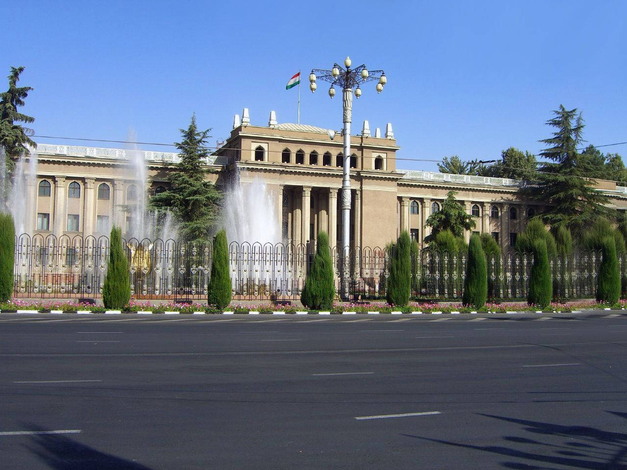 Tajikistan 桌布 壁紙 — 宏偉的新古典主義建築，擁有拱形窗戶和柱廊，矗立在華麗的圍欄和噴泉之後，背景是清澈的藍天。