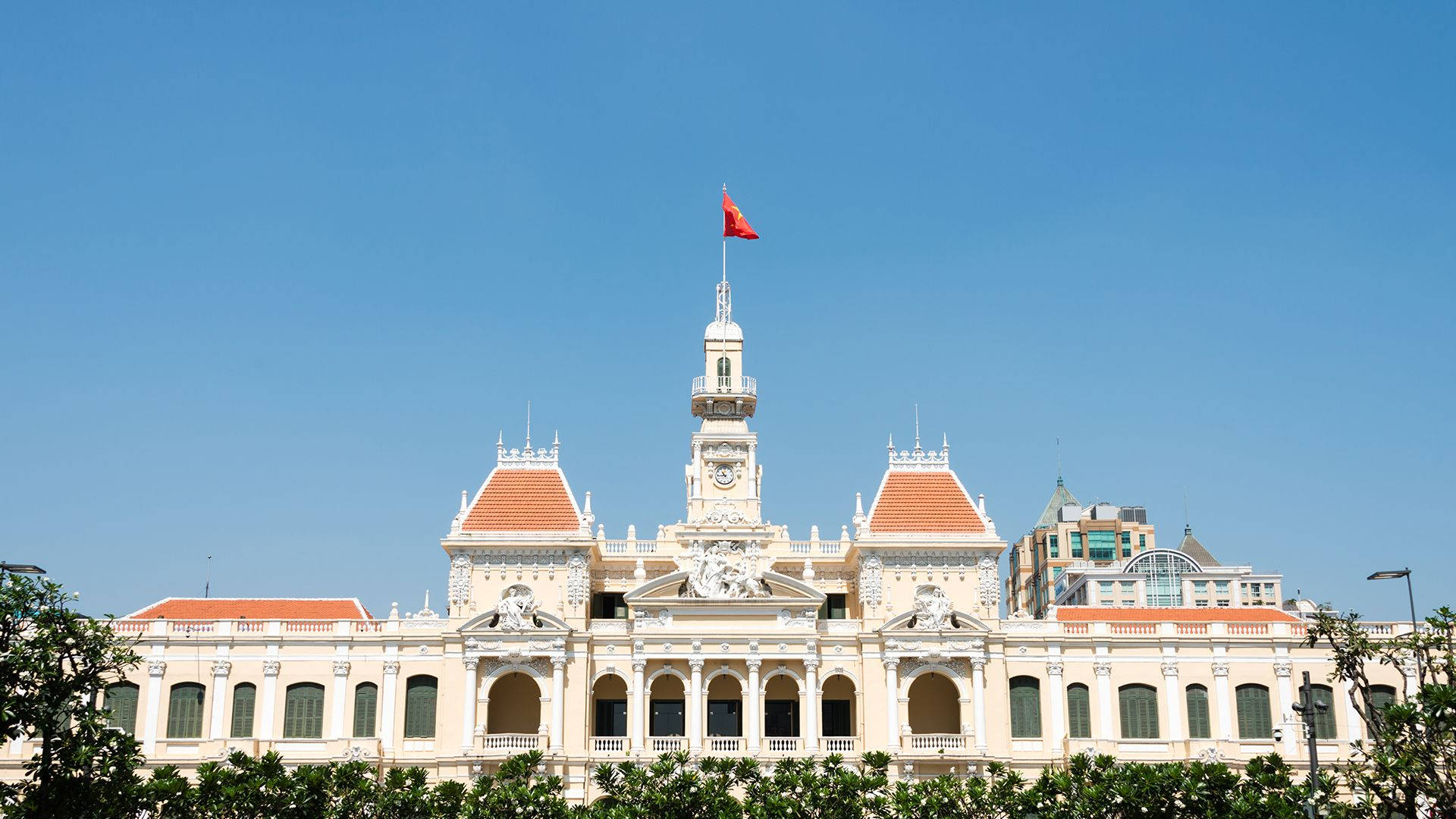 Ho Chi Minh City 桌布 壁紙 — 宏偉的新古典主義建築，擁有紅色瓦片屋頂和鐘樓，在晴朗的藍天下飄揚著紅旗。
