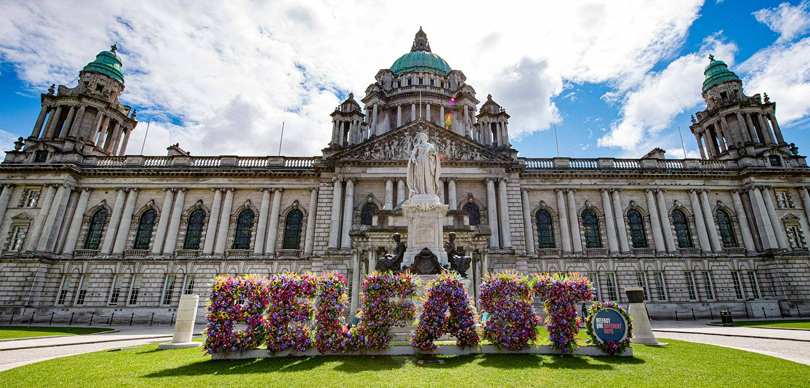 Grass 桌布 壁紙 — 宏偉的新古典主義建築，擁有綠色圓頂和花卉「BELFAST」標誌，在明亮的藍天和蓬鬆的白雲下。