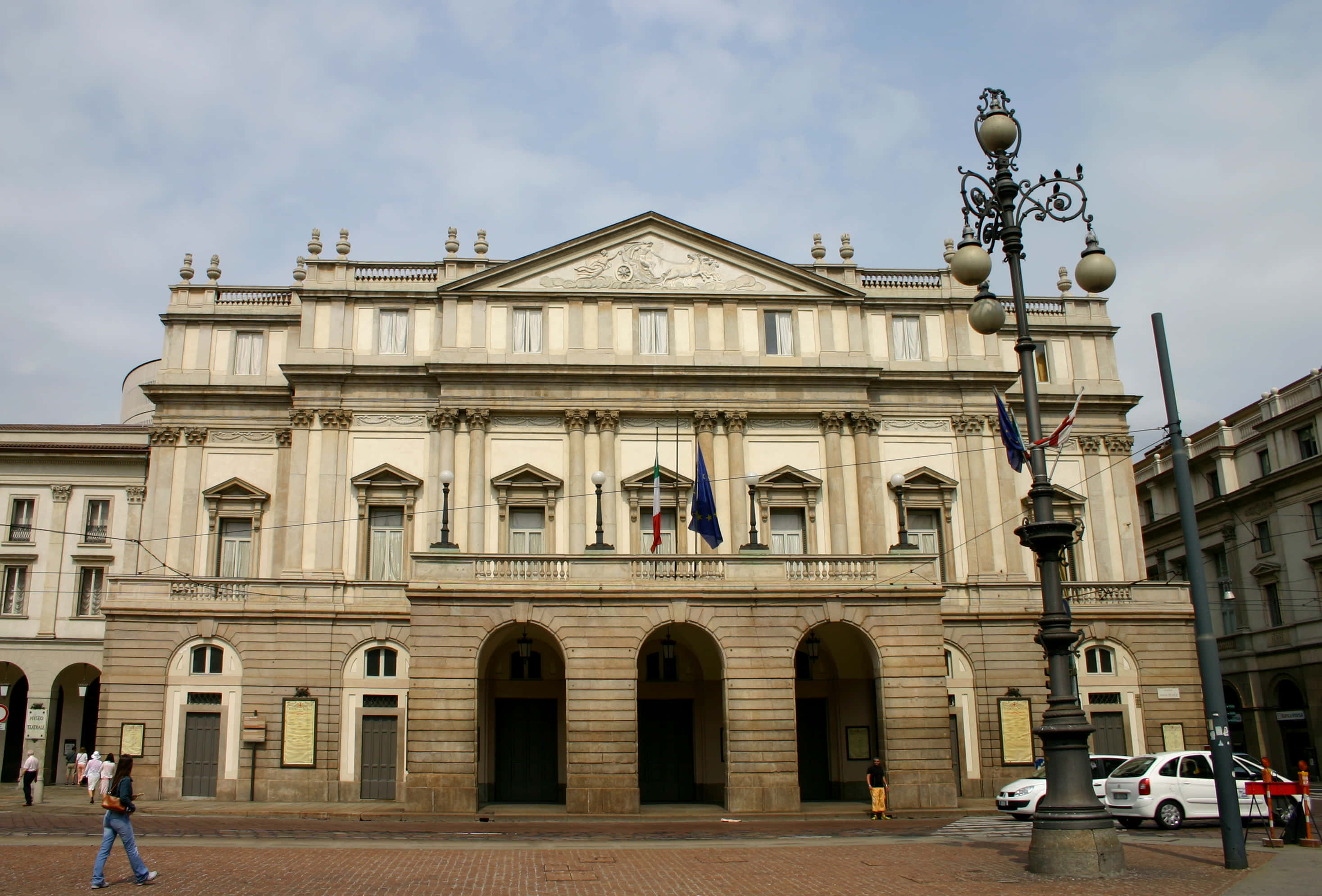 theatre 桌布 壁紙 — 宏偉的新古典主義歌劇院，擁有拱形入口和旗幟，天空多雲。