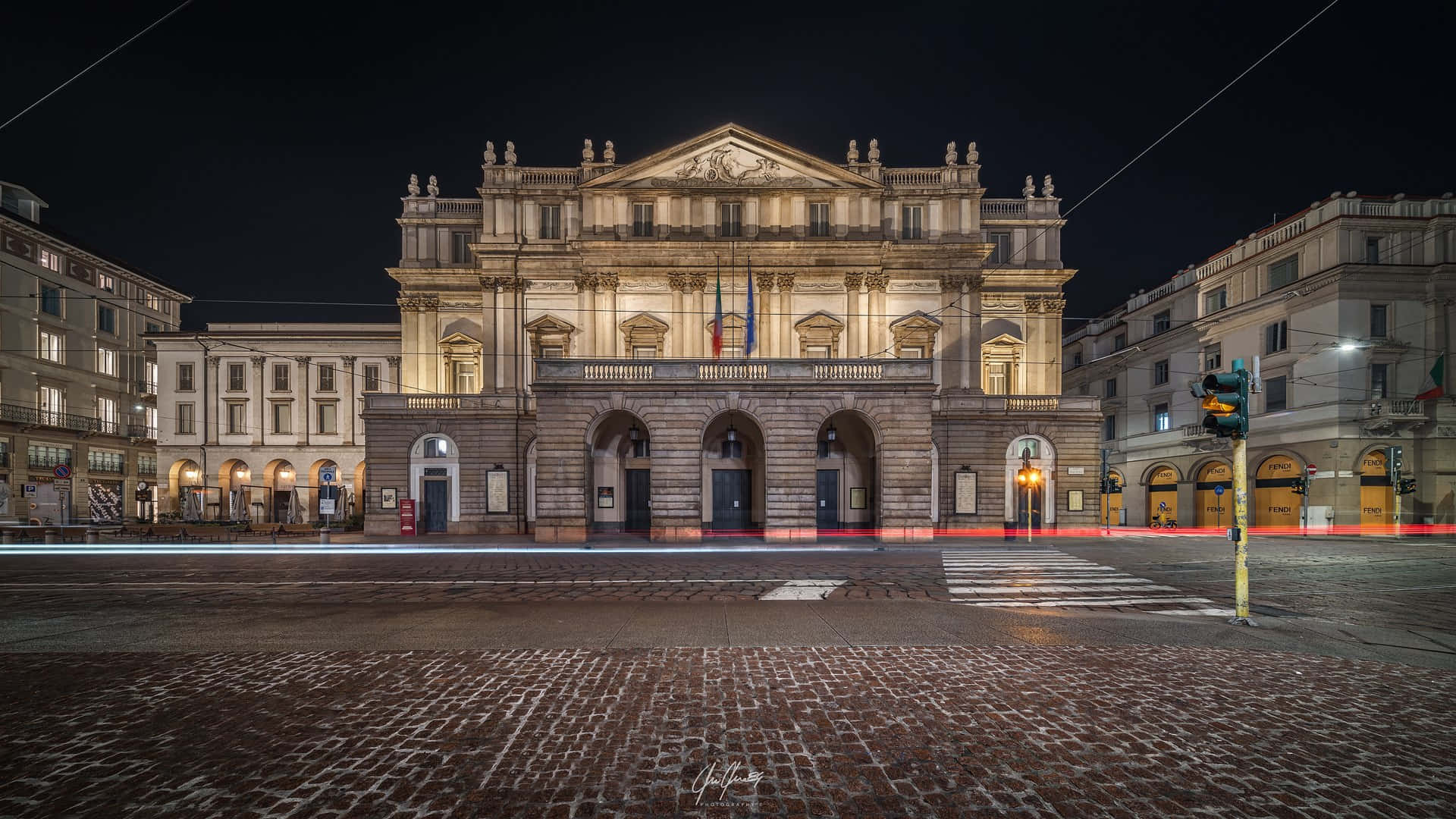 Milan 桌布 壁紙 — 宏偉的新古典主義歌劇院在夜間被照亮，鵝卵石街道上行駛的汽車留下紅色光痕。