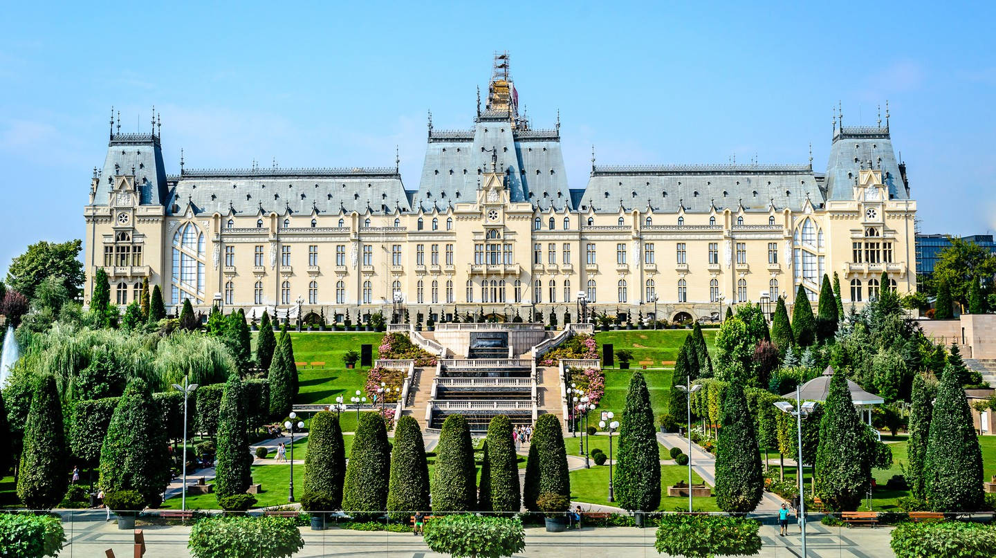 Romania 桌布 壁紙 — 宏偉的新哥德式宮殿，裝飾華麗的塔樓和尖頂，在晴朗的藍天下，雄偉地矗立在修剪整齊的花園後面。
