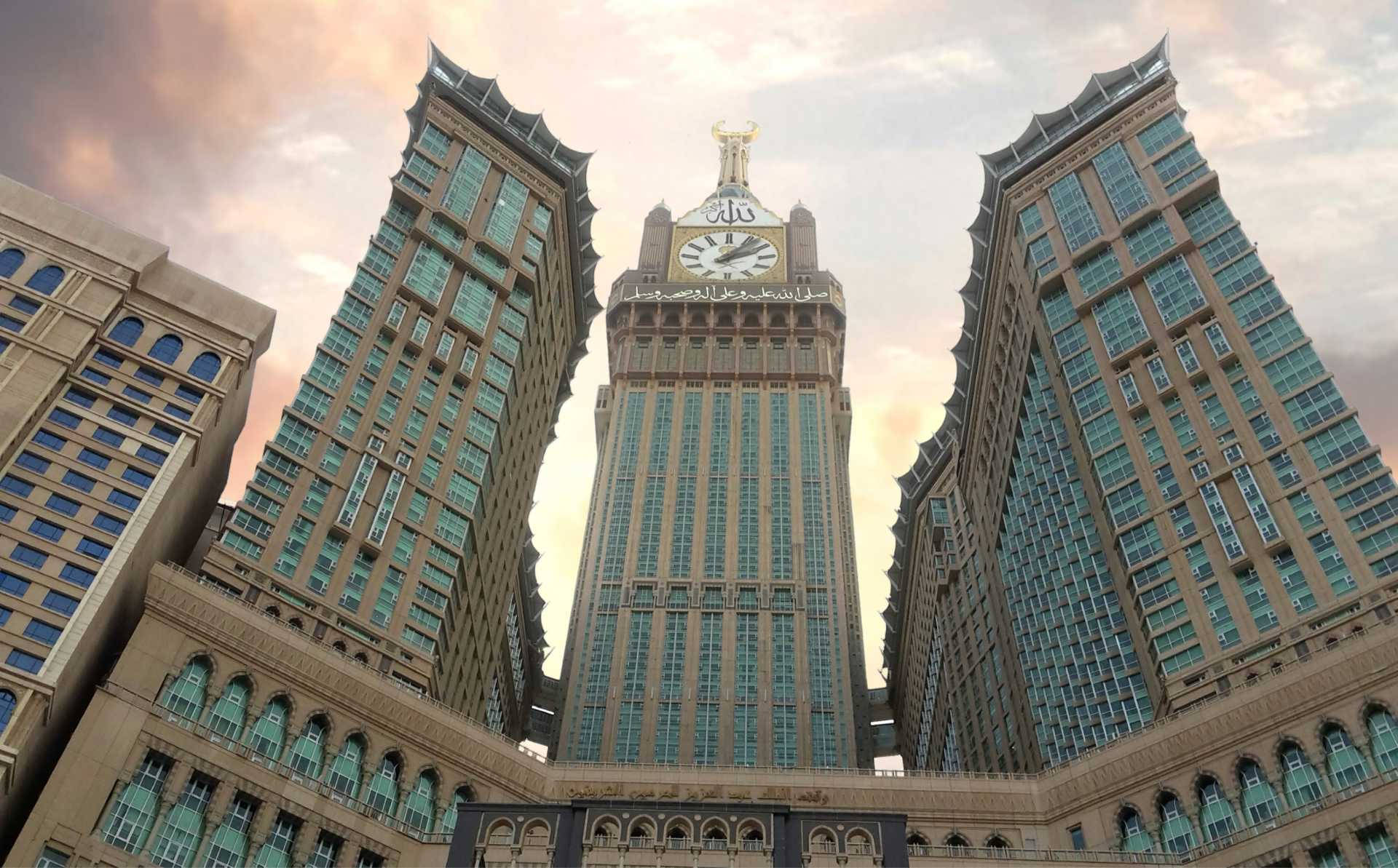 Saudi 桌布 壁紙 — 宏偉的時鐘塔，兩旁是雙子摩天大樓，在柔和夕陽天空和溫暖雲彩下。